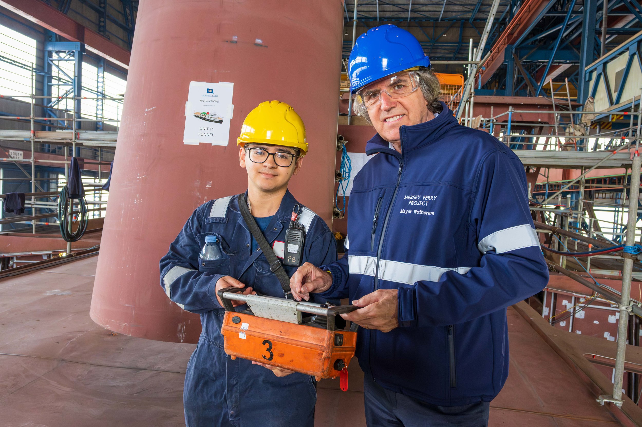 Balaena Engineer in hard hats and safety glasses standing inside a shipyard or construction site, smiling at the camera, surrounded by scaffolding and industrial equipment, with a large ship in the background.
