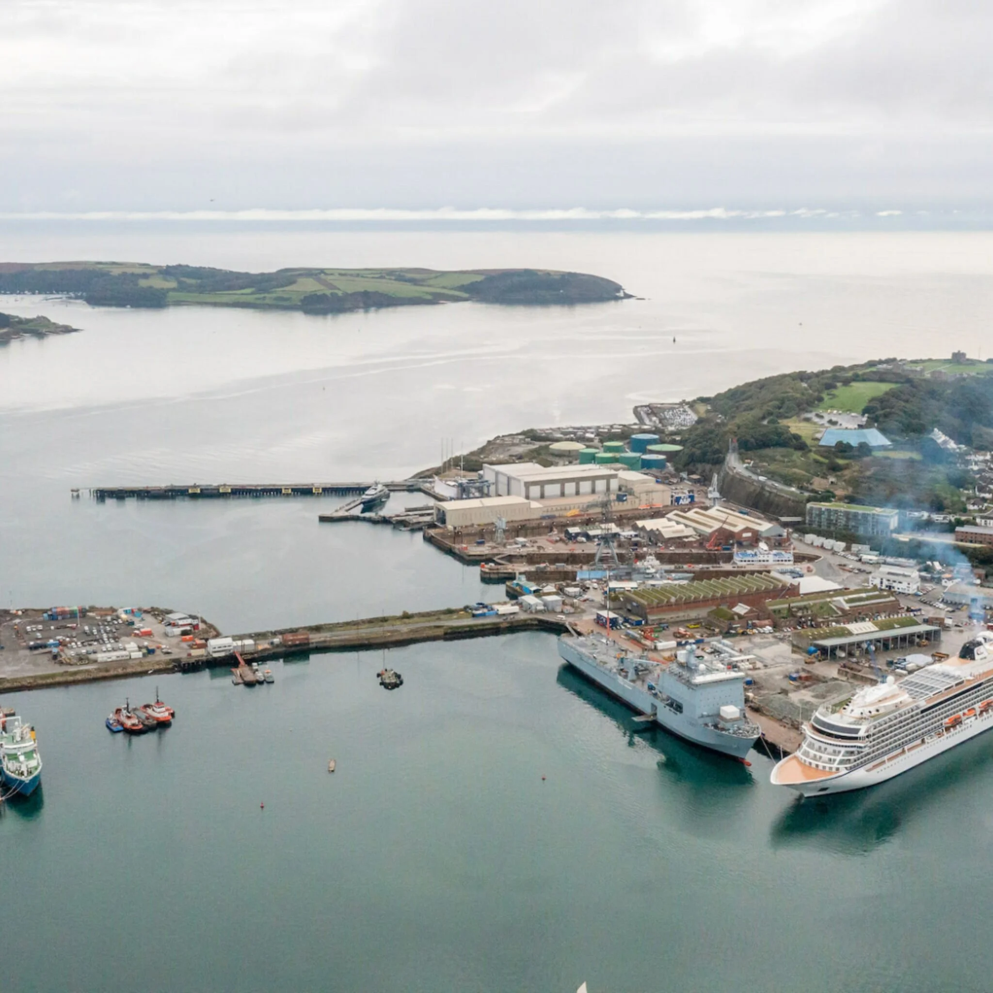 A harbor with boats docked, a pier, industrial buildings, and green hills in the background under a cloudy sky.