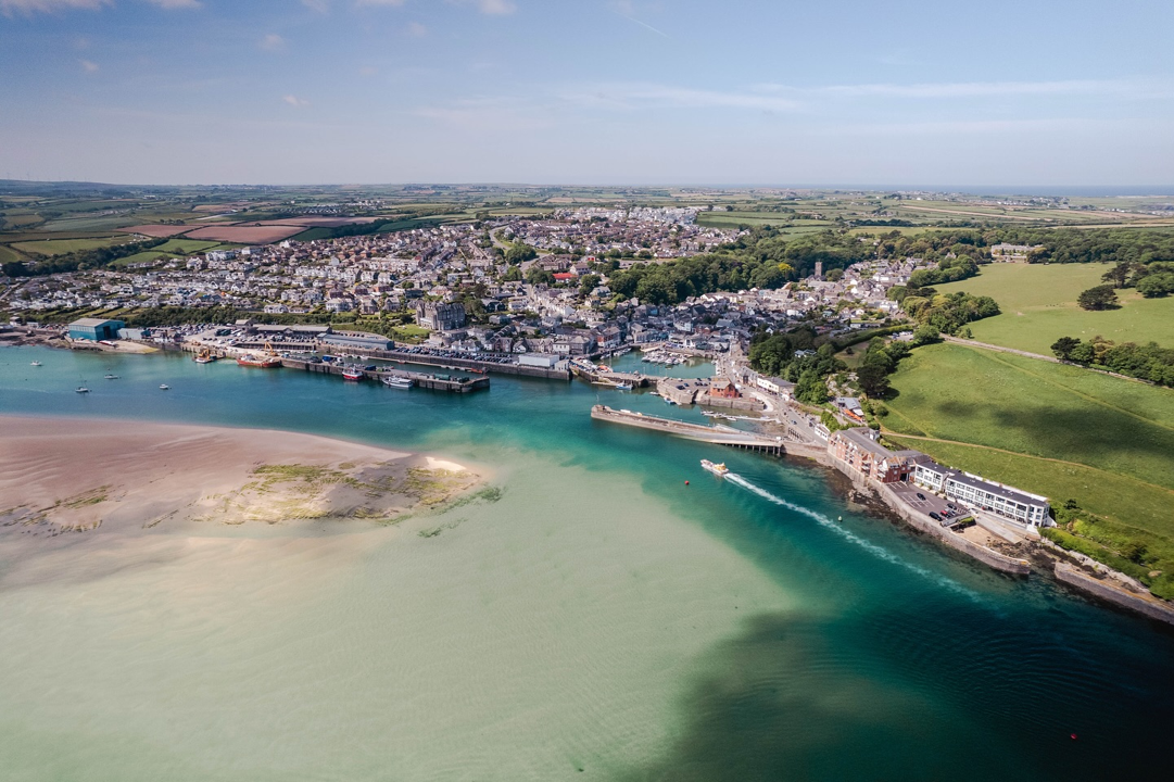 Aerial view of a coastal town with boats in the harbor, sandy beach, and green fields surrounding the area.