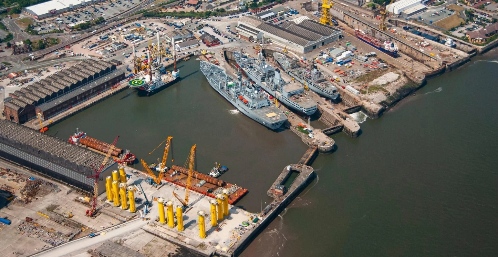 Aerial view of a busy harbor with ships docked, construction cranes, industrial buildings, and surrounding city infrastructure.