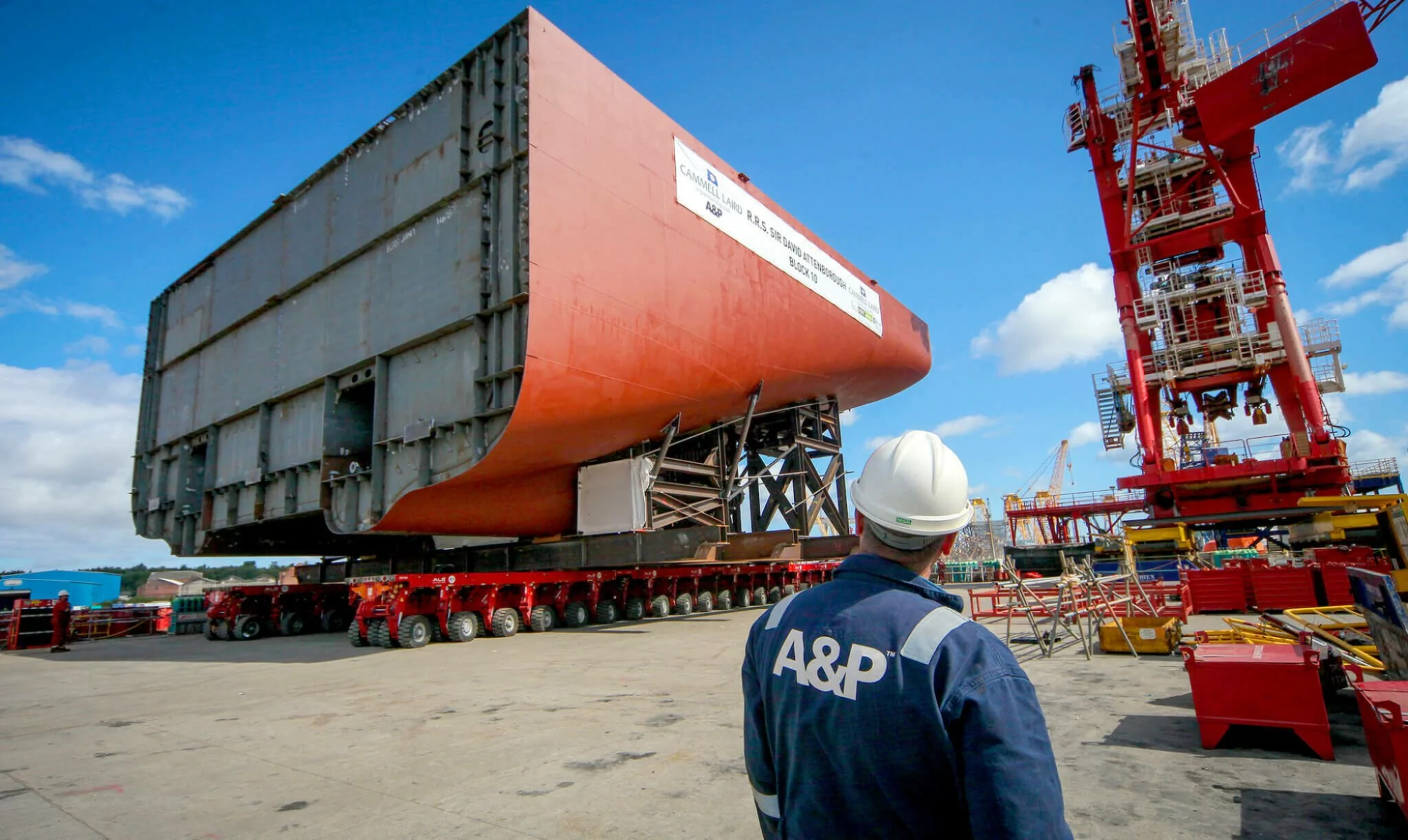 A person wearing a white safety helmet and blue jacket with 'A&P' on the back watching a large cargo structure being transported on a flatbed, with cranes and other industrial equipment in the background on a clear, sunny day.