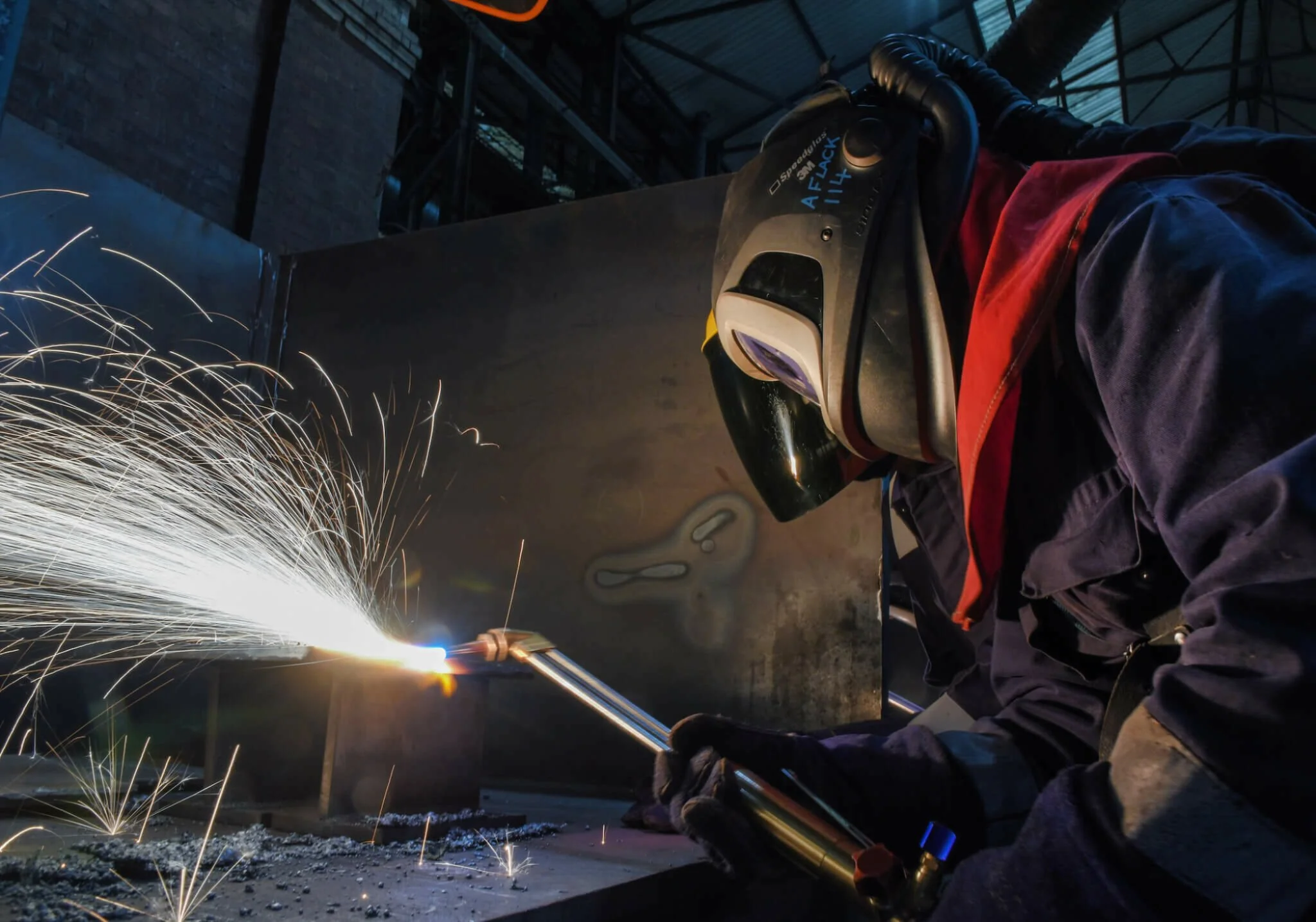A worker welding metal in a factory, wearing protective gear including a welding helmet and gloves, with sparks flying from the welding process.