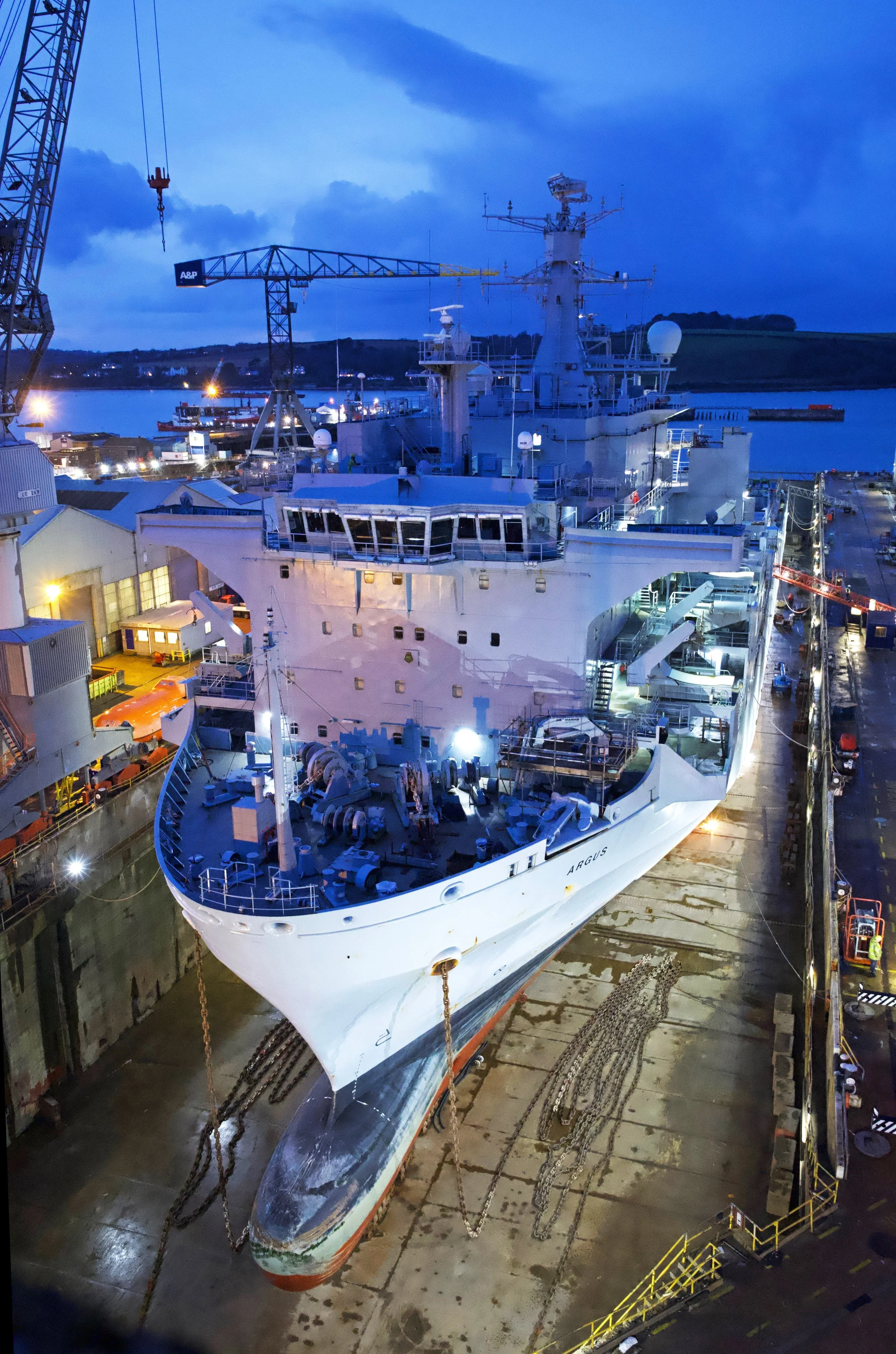 A large naval ship in Balaena Falmouth dry dock under construction or maintenance, with cranes and equipment around, and a body of water and hilly terrain in the background during dusk or dawn.