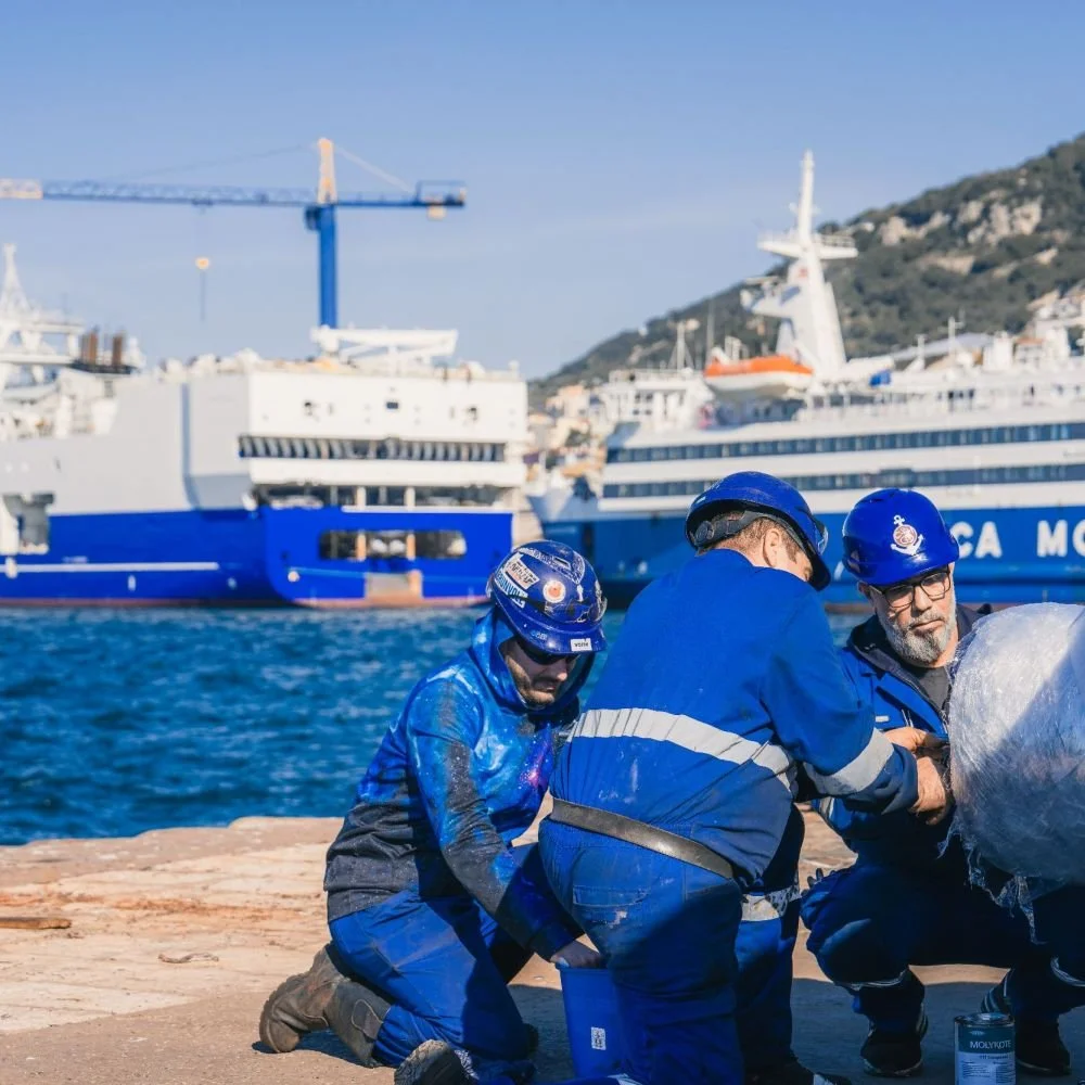 Three crew members in blue uniforms and helmets on a dock working together near the water, with large cruise ships and a hillside in the background.
