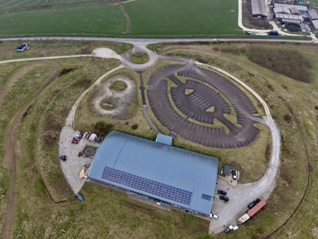 Aerial view of a parking lot and building with solar panels, surrounded by open grassy fields and a rural road.