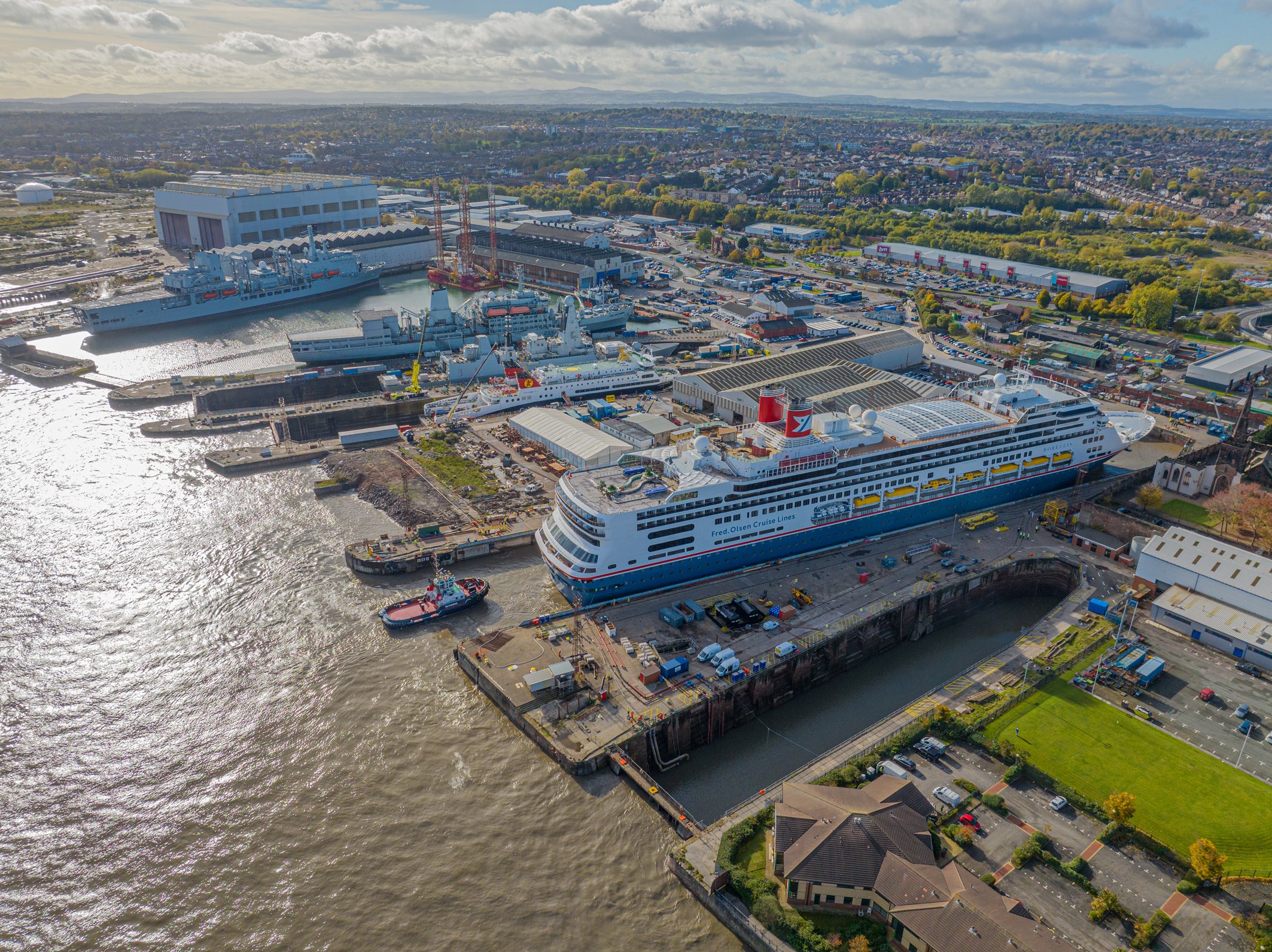 Aerial view of Balaena Birkenhead  with ships, boats, and cranes, adjacent to a residential and commercial area with buildings, parking lots, and greenery under a partly cloudy sky.
