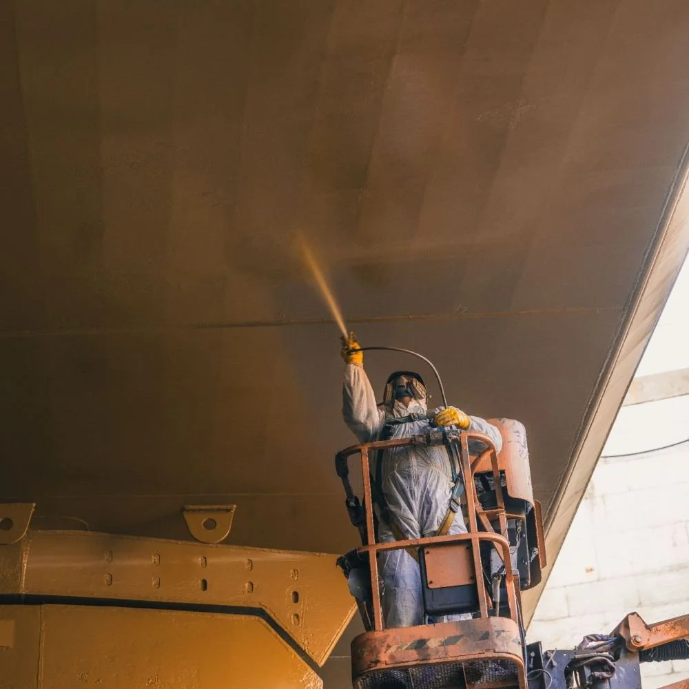 A worker wearing a protective suit, mask, and gloves is on a lift, spray painting or coating the underside of a large structure, possibly an aircraft or industrial equipment.