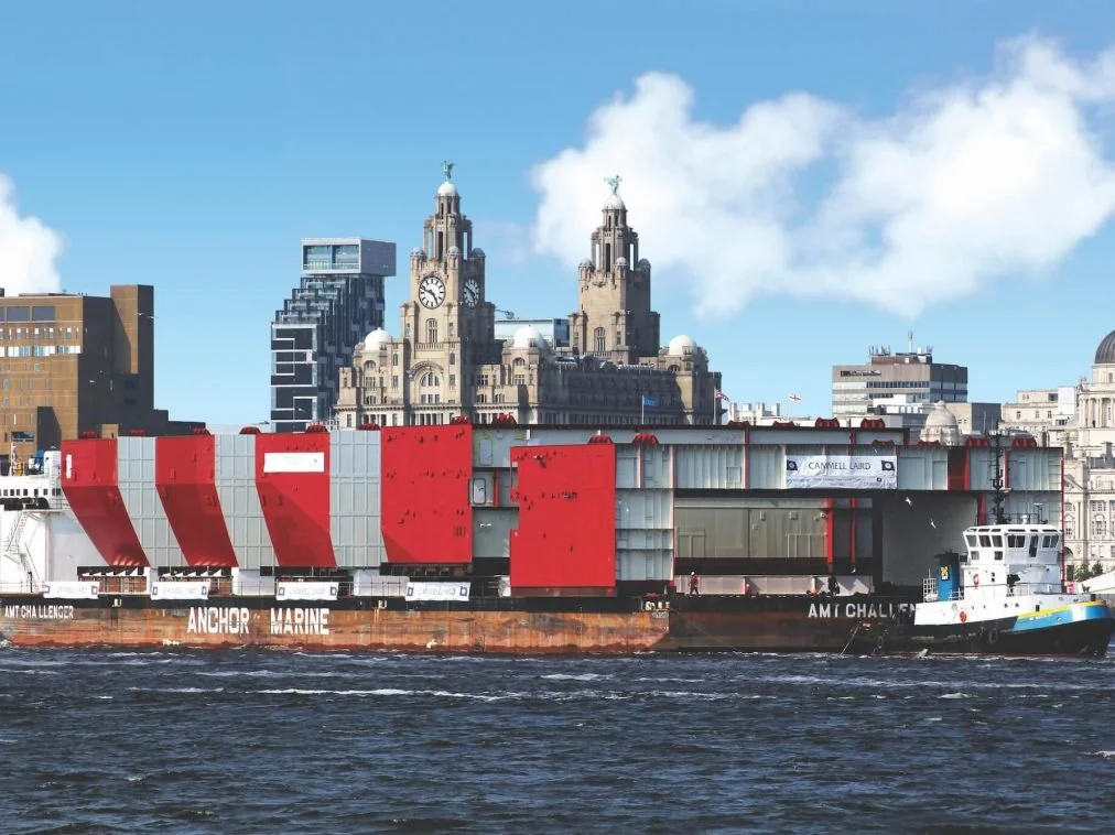 A barge carrying a large structure with white and red sections on a river, with a city skyline featuring a historic building with clock towers and modern buildings in the background.