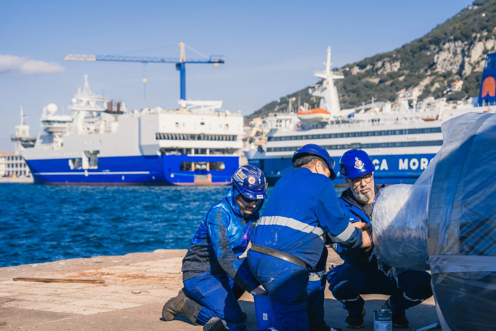 Fishermen repairing a boat on a sandy beach with large cruise ships and a hill in the background.