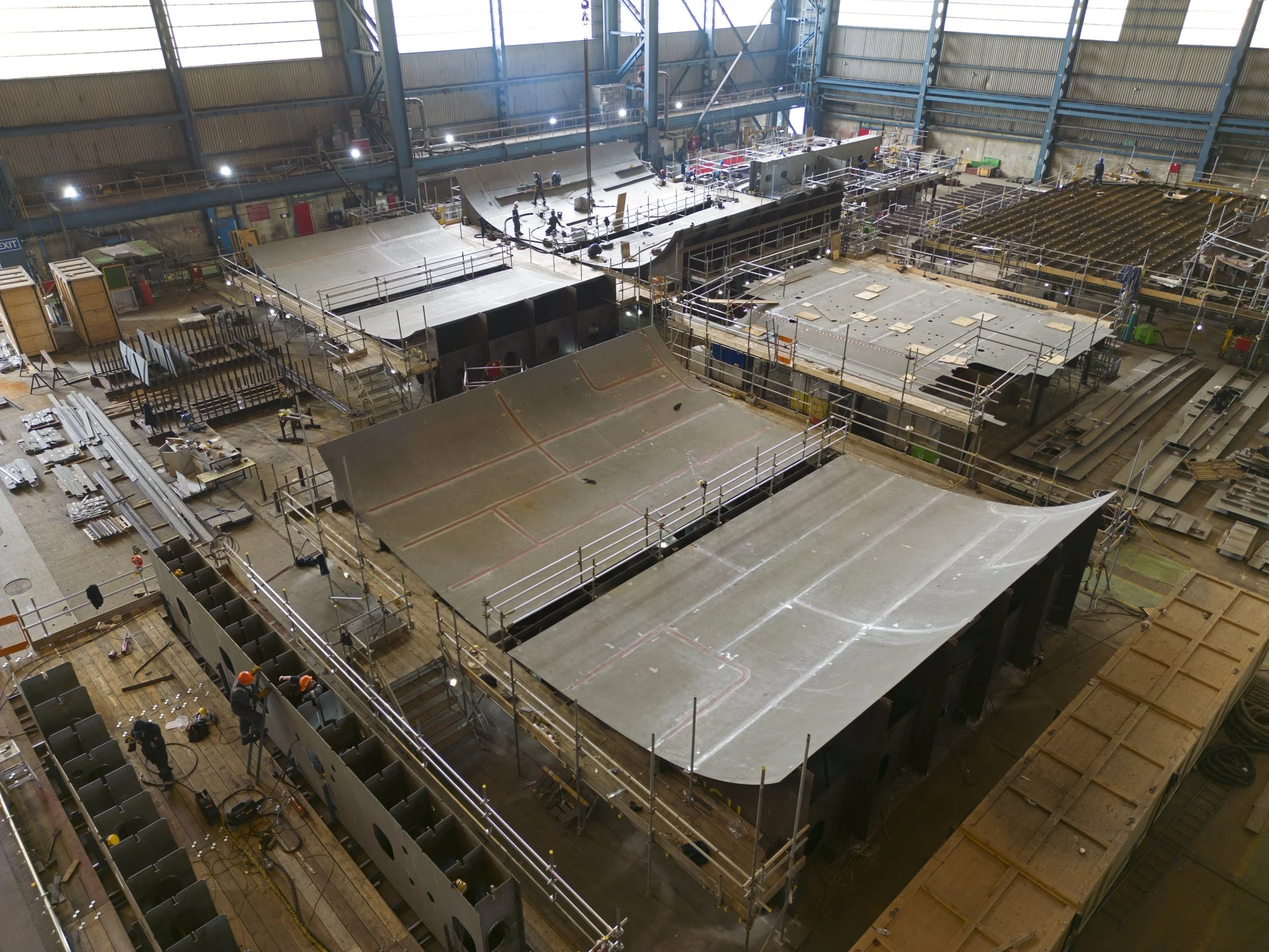 Indoor manufacturing facility in Balena Birkenhead with partially assembled large metal structures, scaffolding, and workers wearing safety helmets.