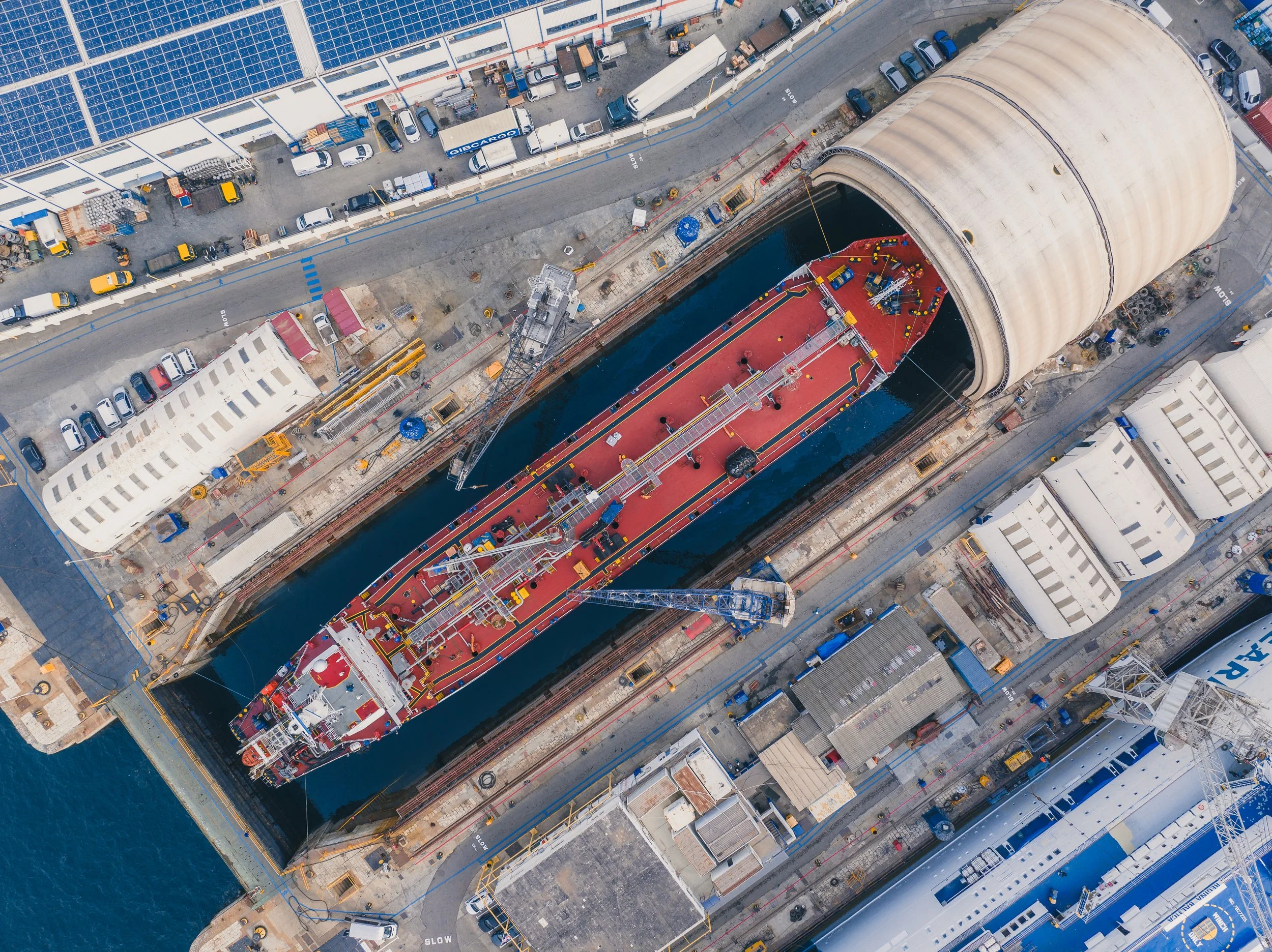 Aerial view of a large cargo ship inside a dry dock with a large cylindrical roof. The ship's deck is red with a crane and equipment, and the dock is surrounded by buildings, parked vehicles, and construction materials.