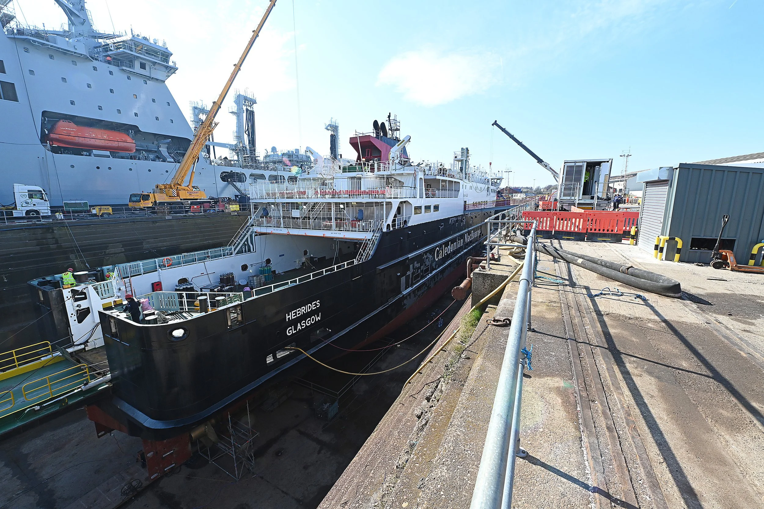 A large passenger and cargo ship docked at a harbor, with construction and maintenance activities underway, including cranes and workers.