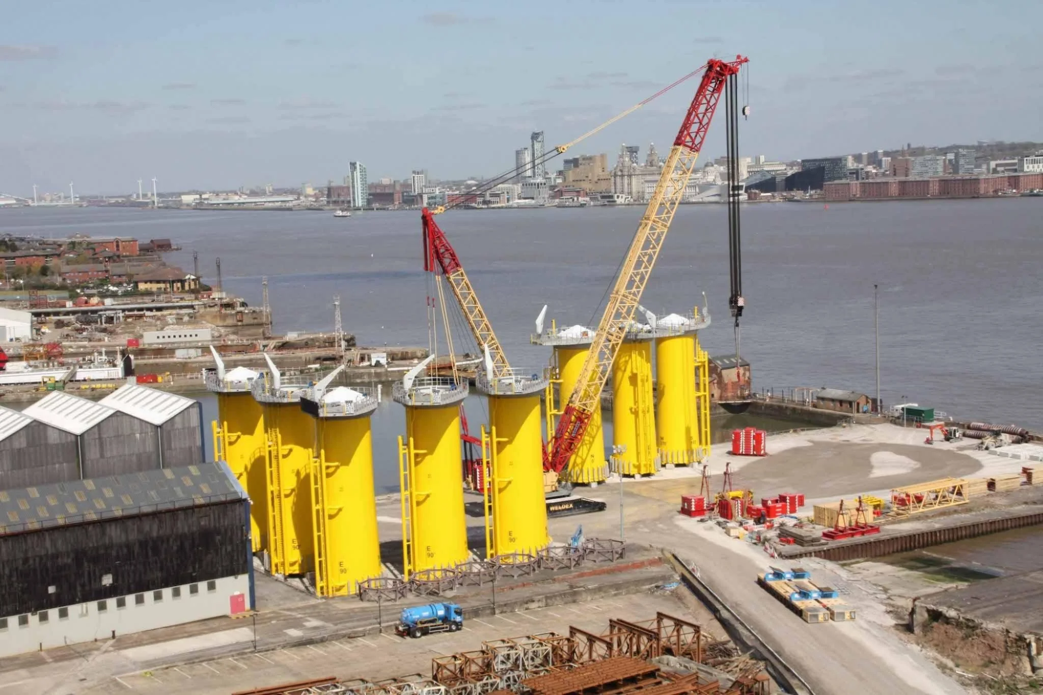 Construction site with large yellow offshore wind turbine foundations and a crane, near a body of water with a city skyline in the background.