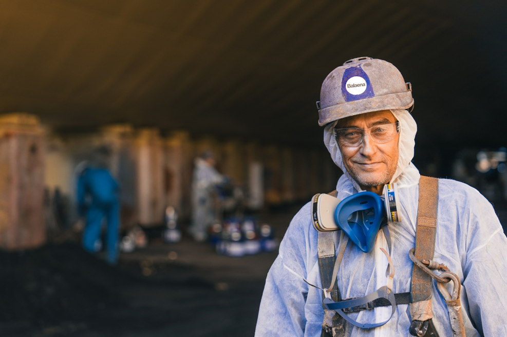 A scientist in protective gear and helmet smiles at the camera, with coworkers working on a large underground excavation site in the background.