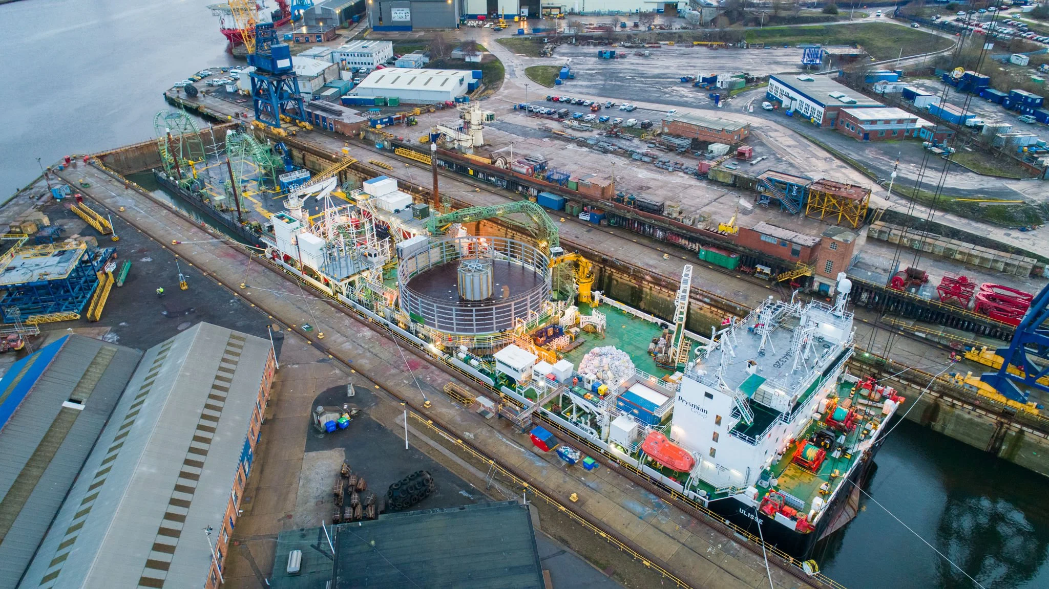 Aerial view of a busy harbor with a large ship docked, containing amusement park rides and structures. Surrounding the harbor are industrial buildings, parking lots, and roads with cars.