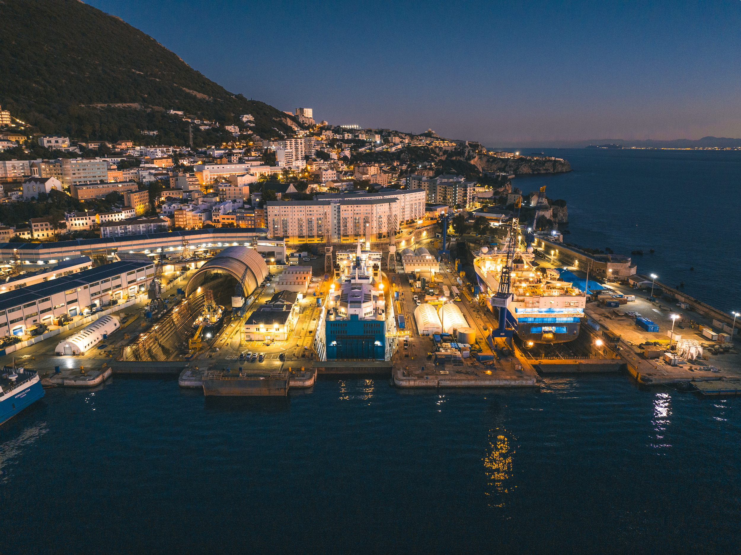 Nighttime view of a harbor with illuminated buildings along the shoreline and a hillside town in the background, reflecting in the water.
