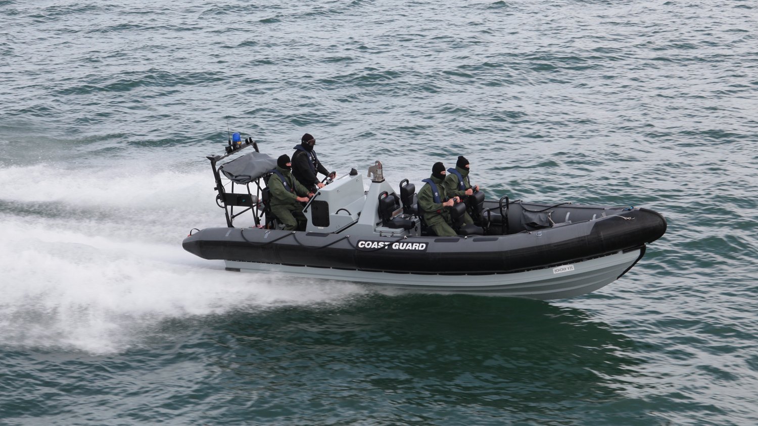 Coast guard members on a black and gray inflatable boat with the word 'COAST GUARD' on the side, patrolling on the water.