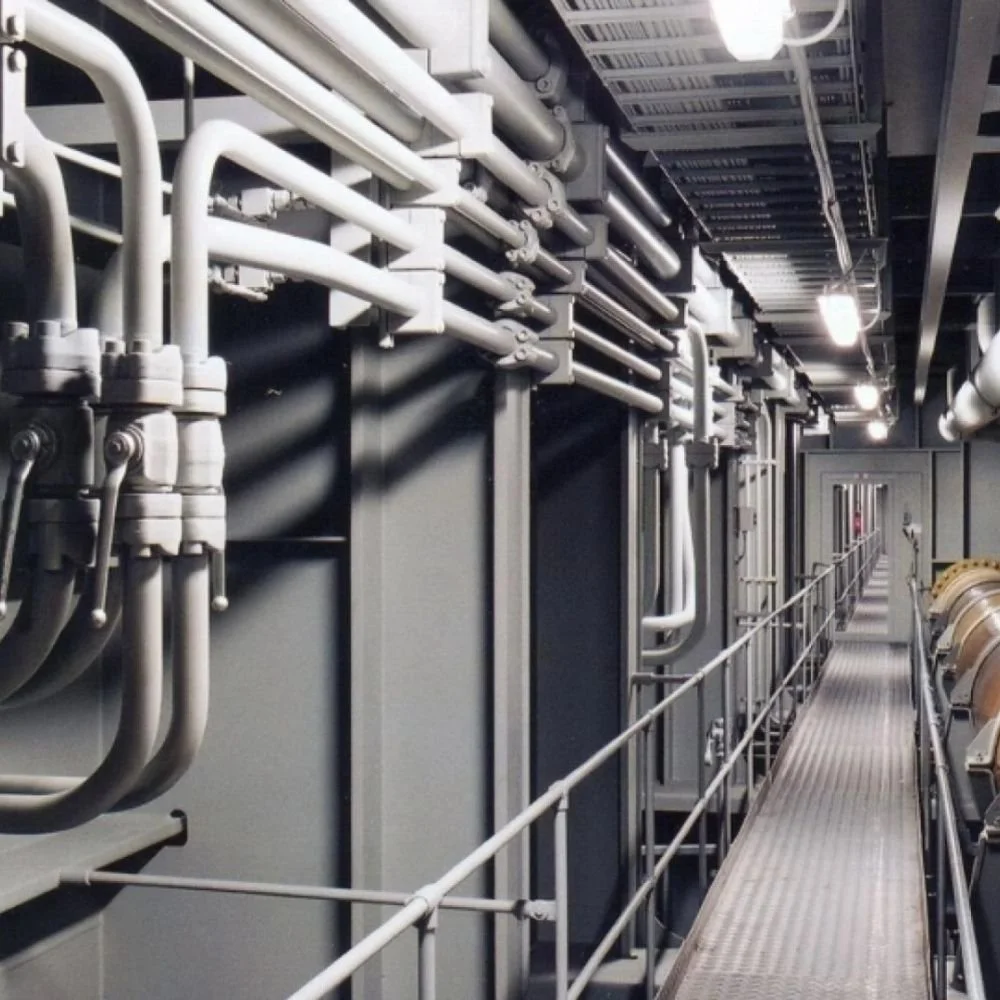 An industrial corridor with metal pipes and ducts running along the ceiling and walls, with a metal walkway and railing for maintenance access.