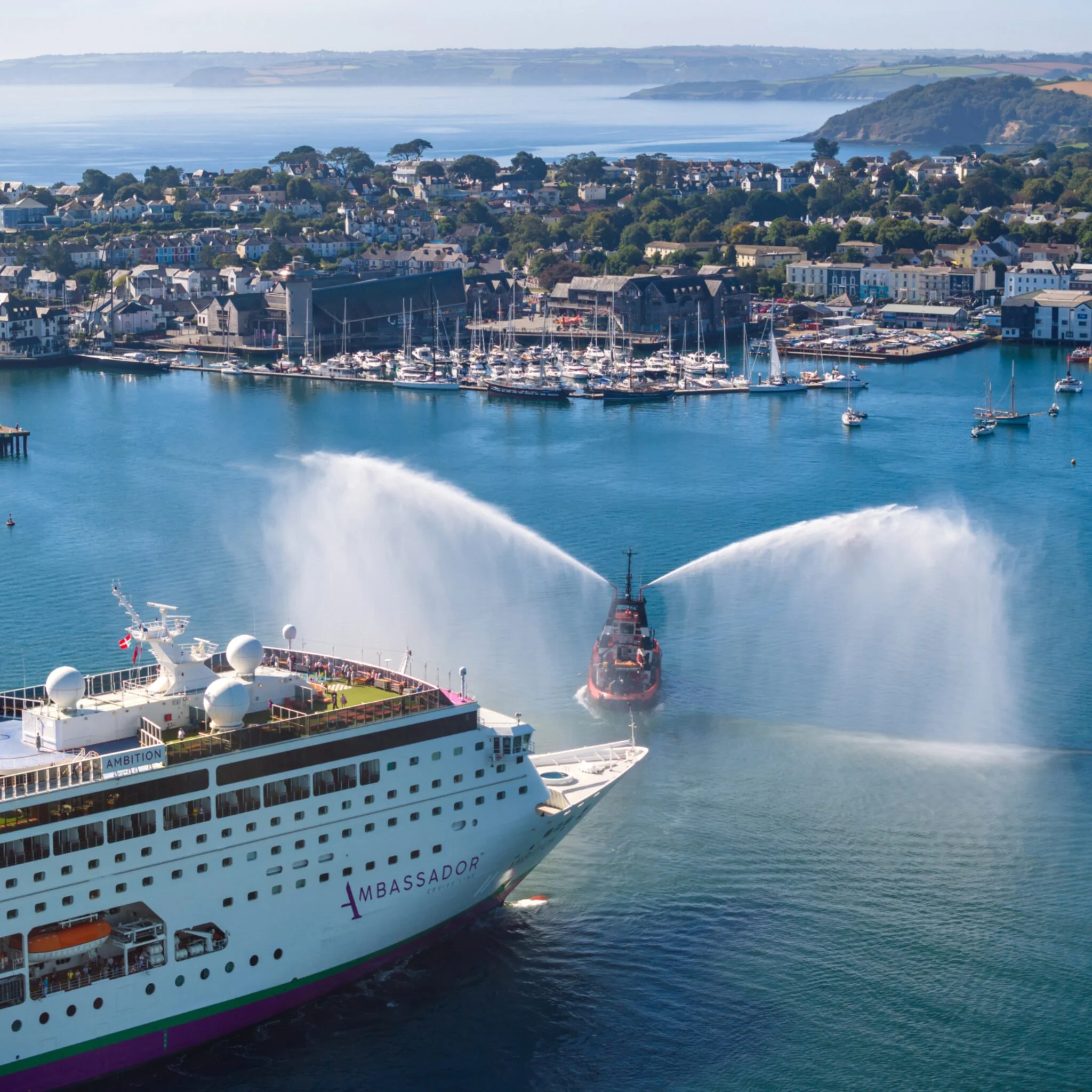A large cruise ship named 'Ambassador' in a harbor, with water spray from a rescue boat approaching. The harbor is filled with boats, and the shore has a town with houses and buildings, backed by green hills and water bodies.