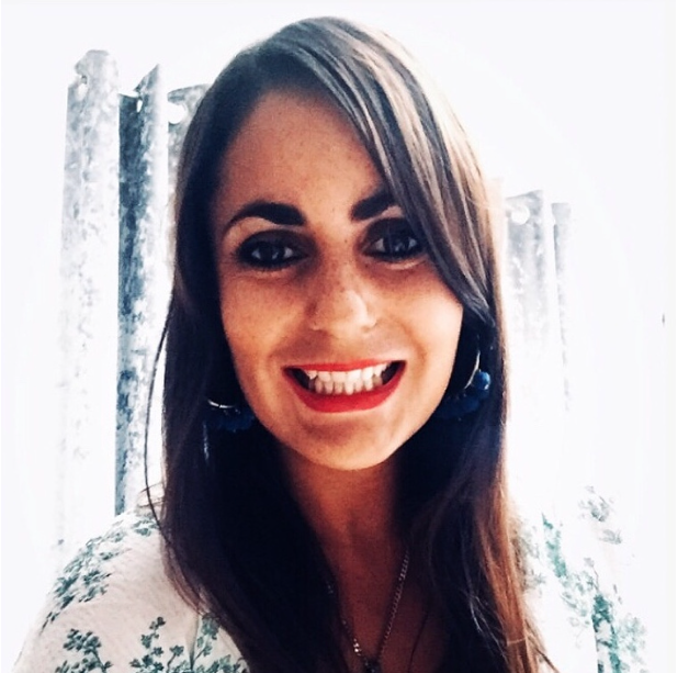 Young woman with long dark hair, wearing red lipstick, earrings, and a floral top, smiling in front of a curtain with a snowy winter scene visible outside.