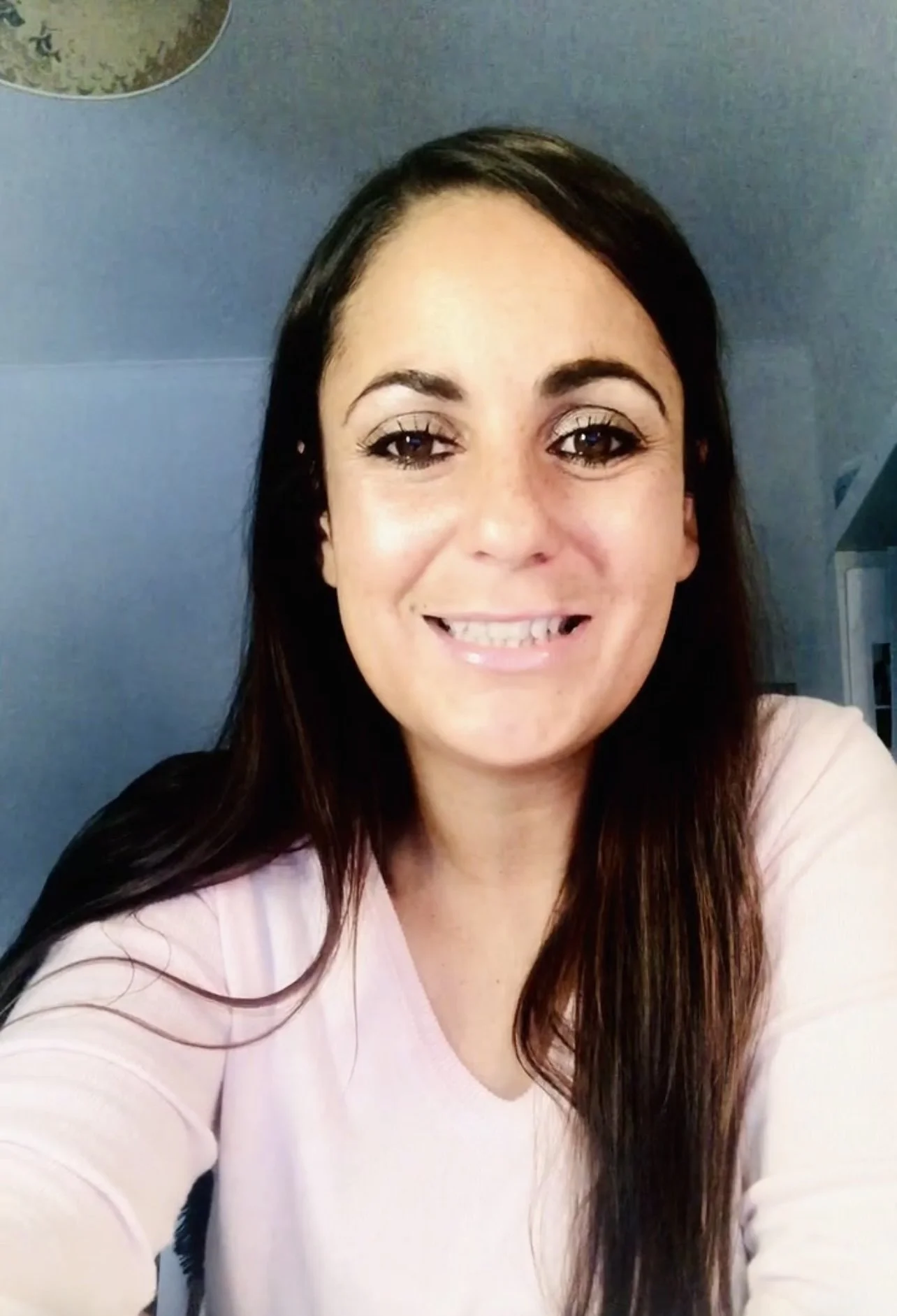 A smiling woman with long dark hair, wearing a light pink top, taking a selfie indoors against a blue wall.
