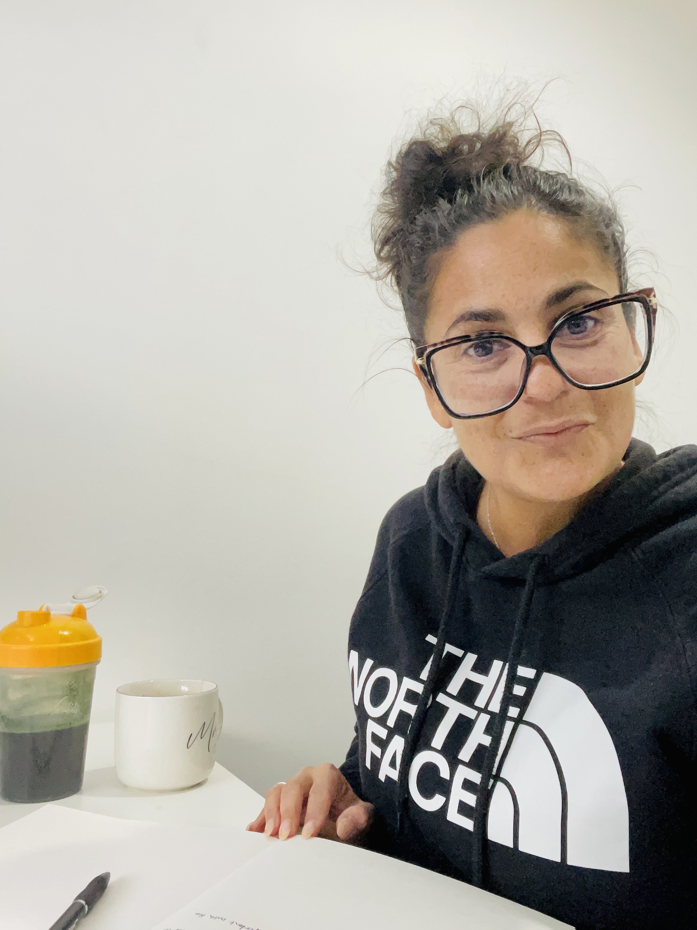 A woman with curly hair tied in a bun and glasses, wearing a black North Face hoodie, sitting at a desk with a white wall background. On the desk, there is a white mug, a book or notebook, a pen, a jar of green tea with an orange lid, and some papers.