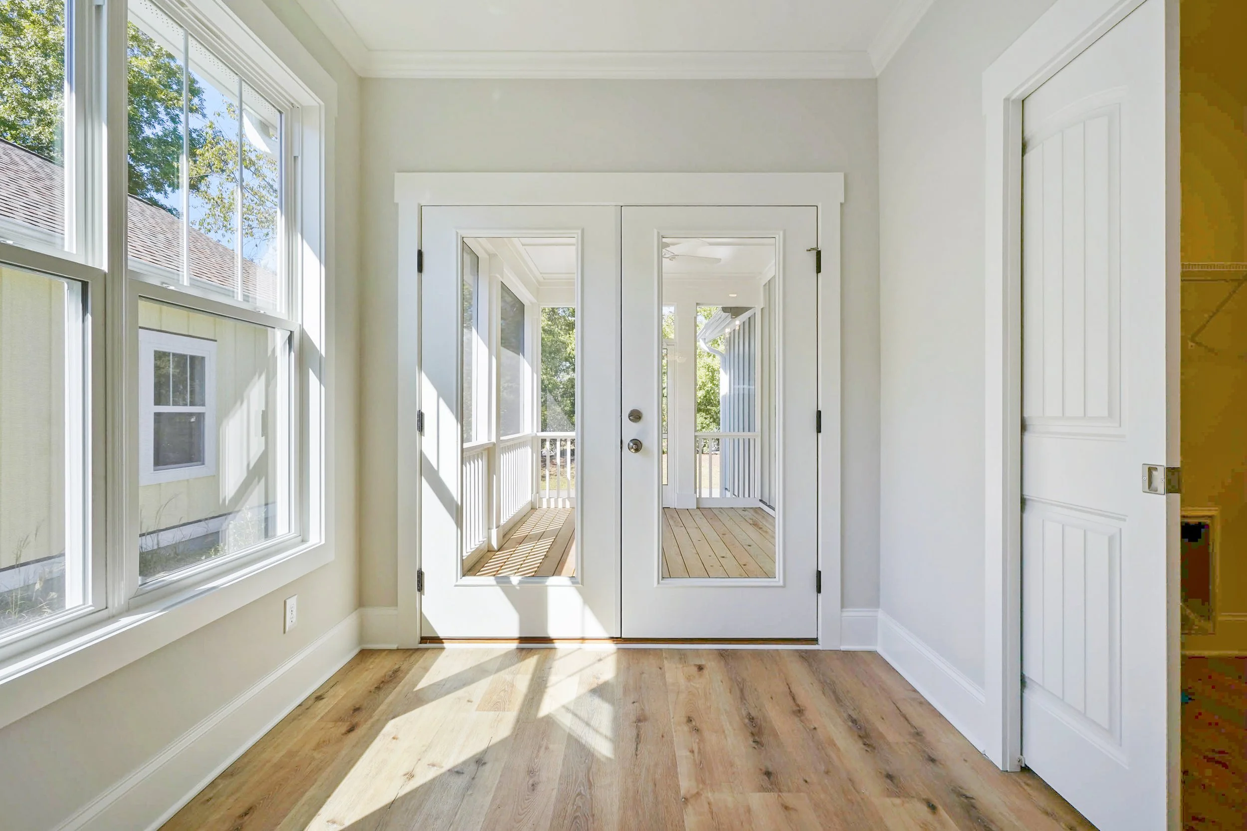 Bright interior room with large windows, white walls, and a wooden floor, leading to a screened porch with a wooden deck outside.