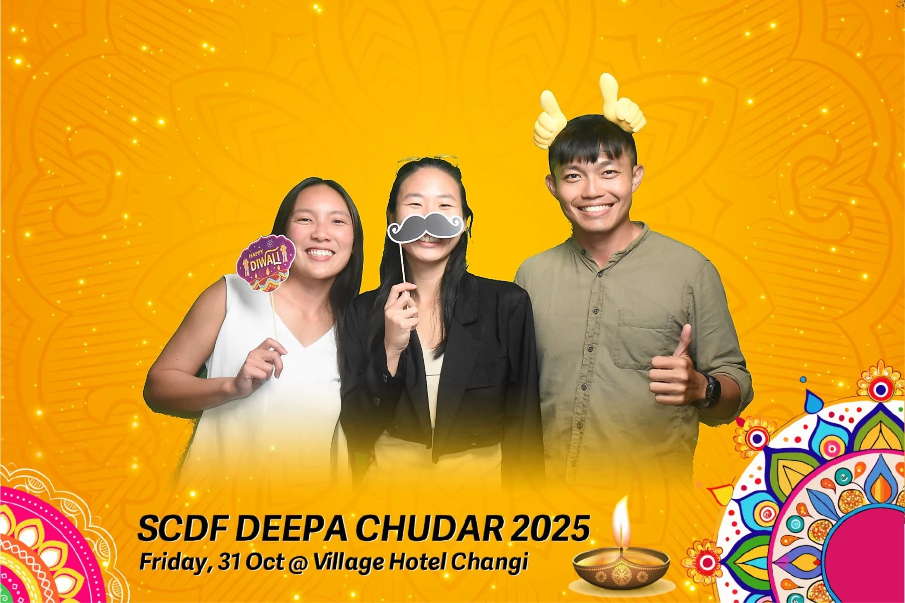 Three people standing together at an Diwali celebration, holding festive signs and wearing fun photo props, with a bright yellow ornate background and traditional Diwali motifs.