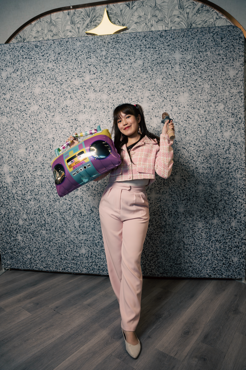 A woman smiling and posing with a microphone in one hand and a colorful boombox-shaped balloon in the other, standing in front of a textured gray backdrop.