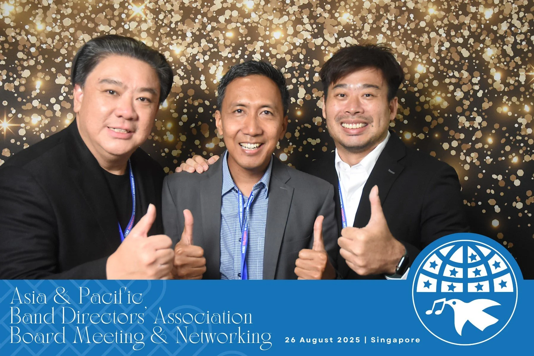 Three men in business attire smiling at the camera and giving thumbs-up gestures at the Asia & Pacific Band Directors' Association Board Meeting & Networking event, in Singapore, on August 26, 2025. The background features sparkling gold and black co