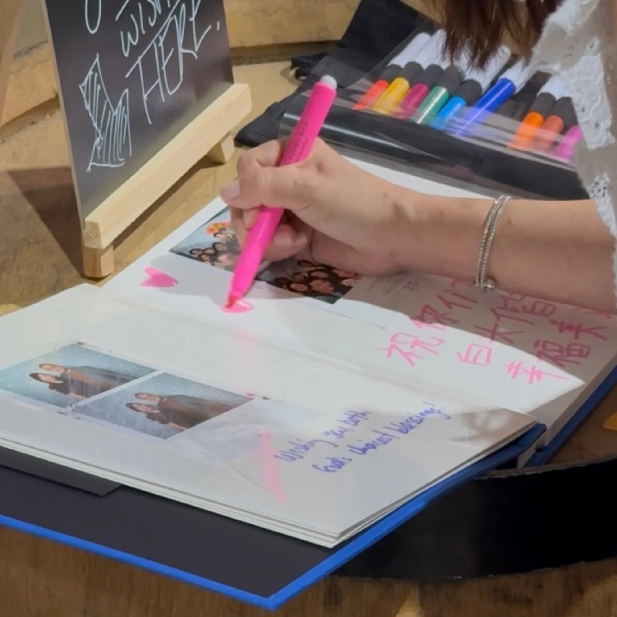 A person writing in a scrapbook with pink marker, adding hearts and Chinese characters. There are photo stickers and handwritten messages, and a set of colorful markers on the table.