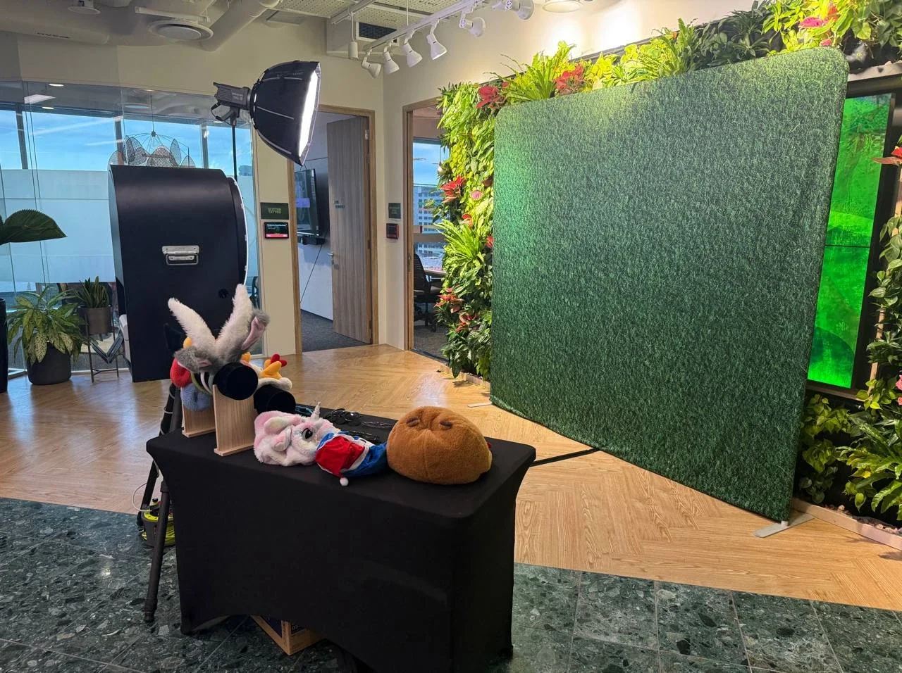 Indoor photo of a photo booth setup with a green divider, plush toys on a black table, and professional lighting equipment. Potted plants and a glass window are visible in the background.