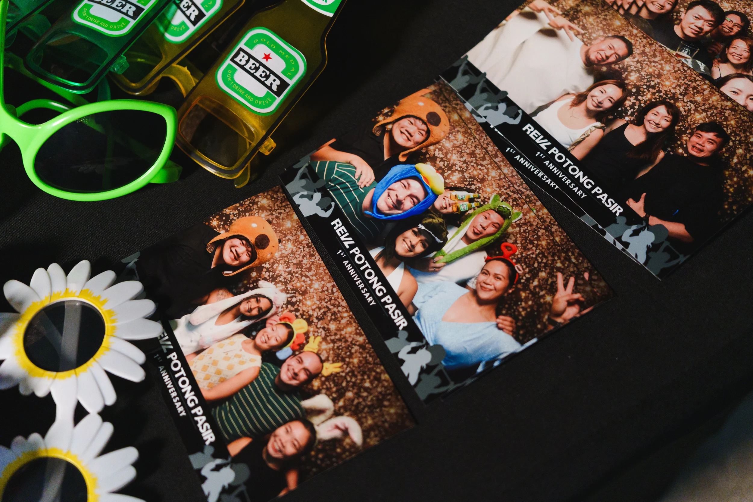 Photobooth strip with multiple pictures of smiling people in colorful costumes and accessories for a 1st anniversary celebration at Revel Potong Pasir, with glasses and decorative items visible in the scene.