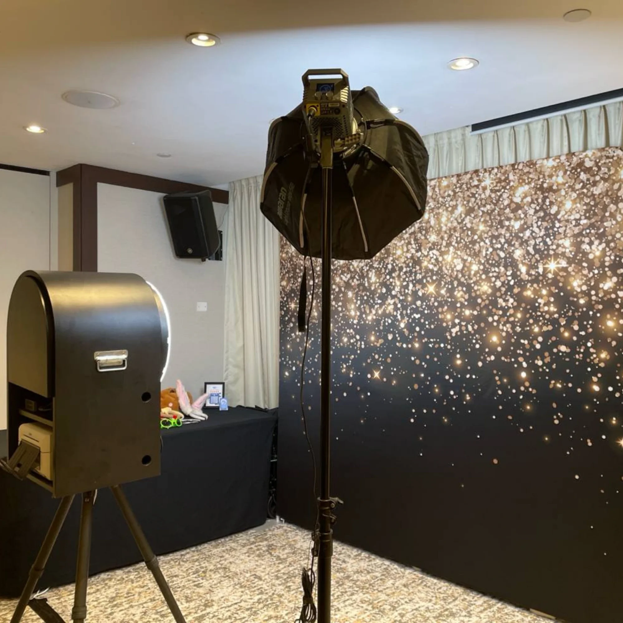 Photo of a photo booth setup with a backdrop featuring a pattern of gold and white dots resembling stars, a professional lighting umbrella, and a camera mounted on a tripod.
