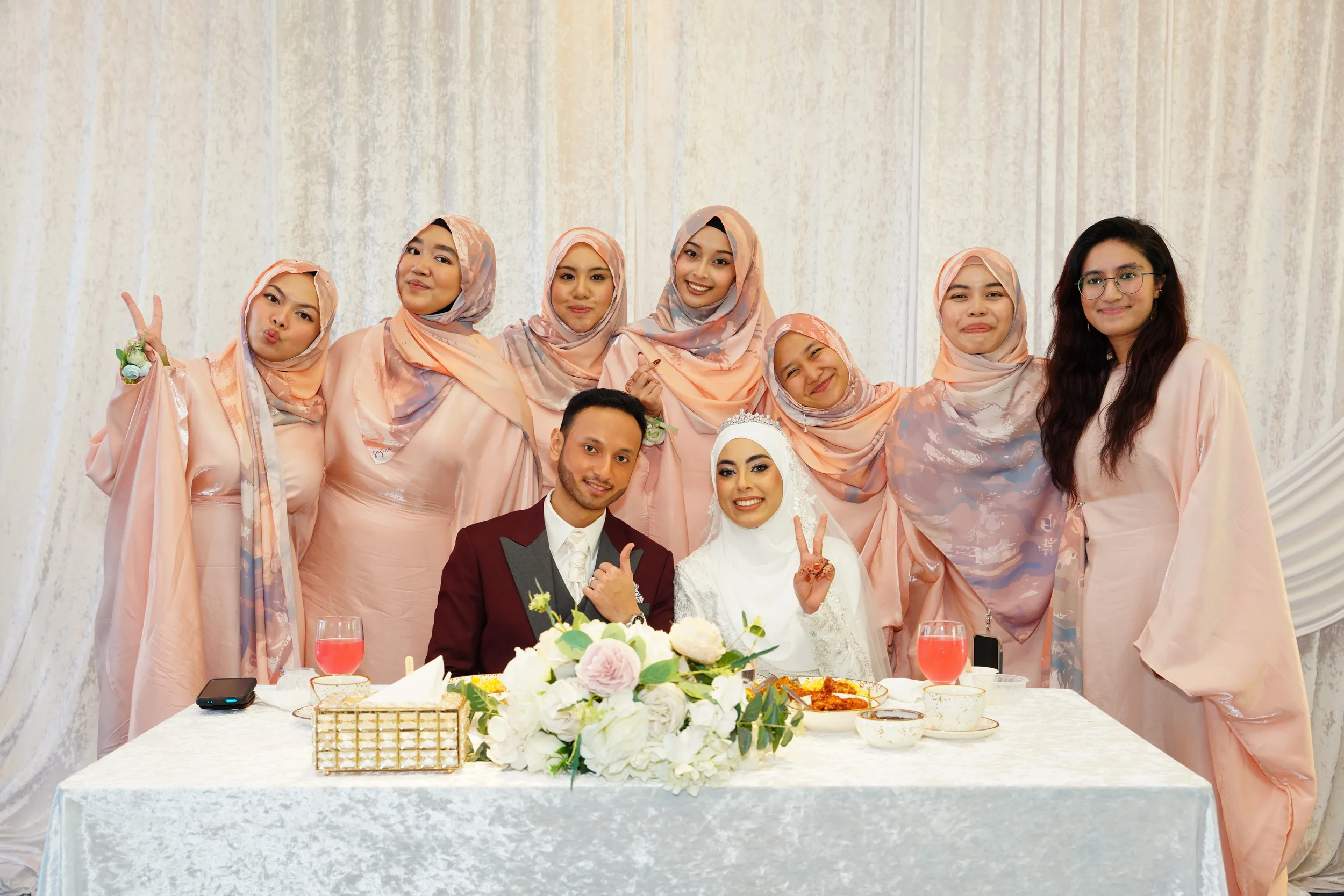 A wedding celebration with a bride and groom seated at a table with friends and family. The bride is wearing a white hijab and dress, making a peace sign. The groom is in a maroon tuxedo. The group is smiling, with some making peace signs or playful gestures, and they are all dressed in pastel pink attire, standing against a light-colored curtain backdrop. There are flowers and food on the table.