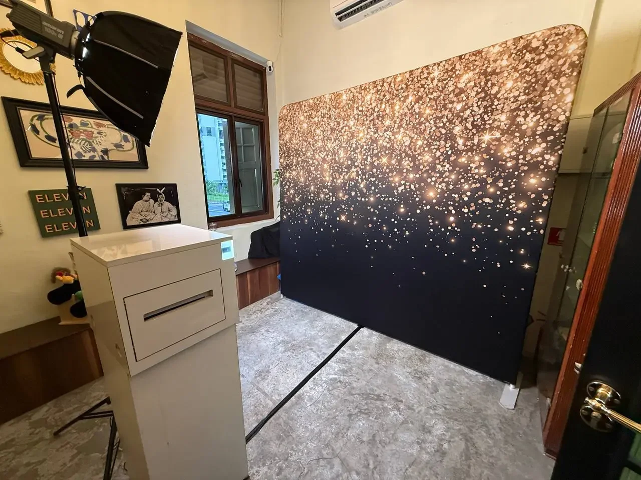 A photography studio setup with professional lighting, a large backdrop with a sparkling gold and black design, a window, wall art, and furniture.