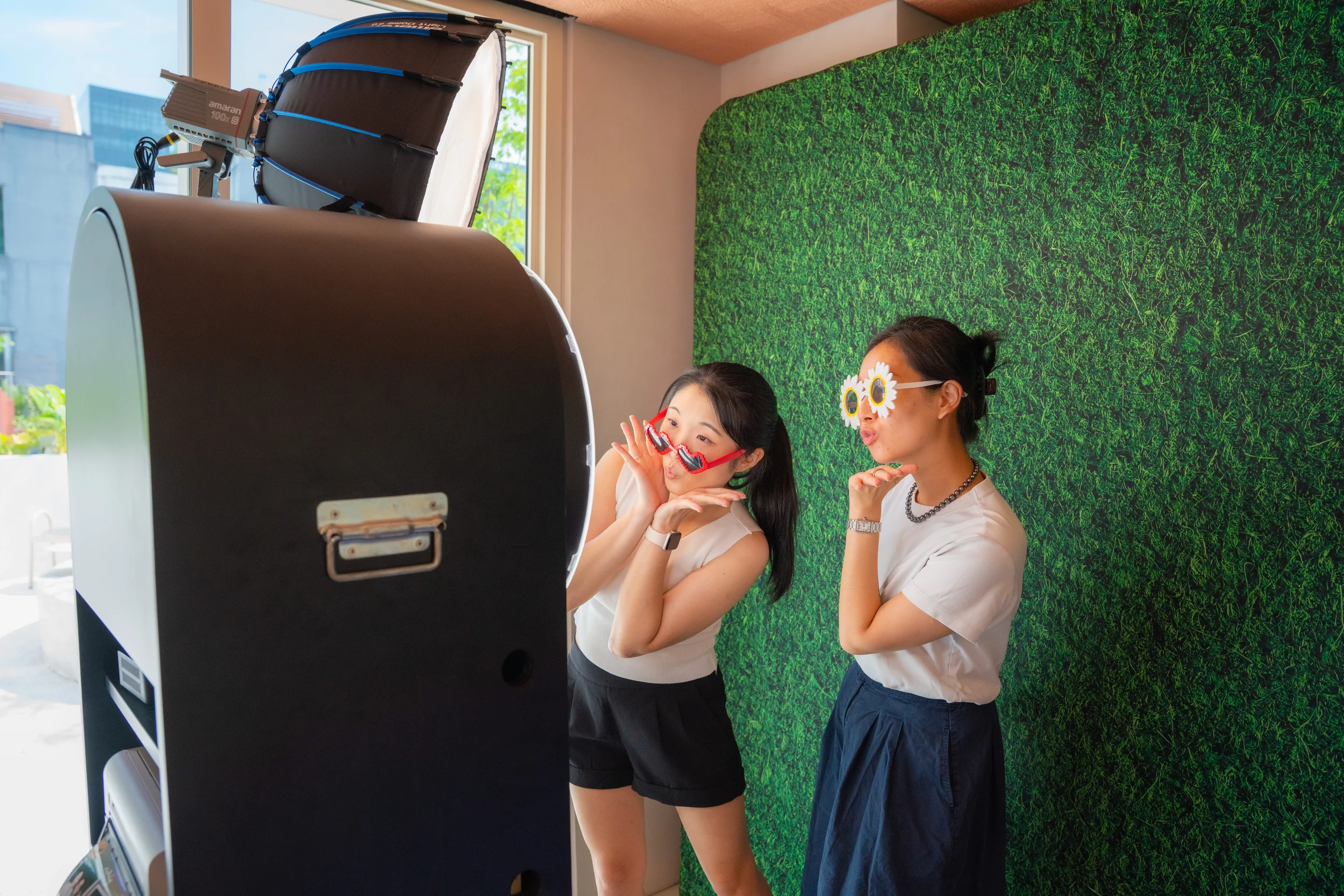 Two women with playful sunglasses and sunglasses accessory posing in front of a photo booth with a green textured backdrop and a small window in the background.