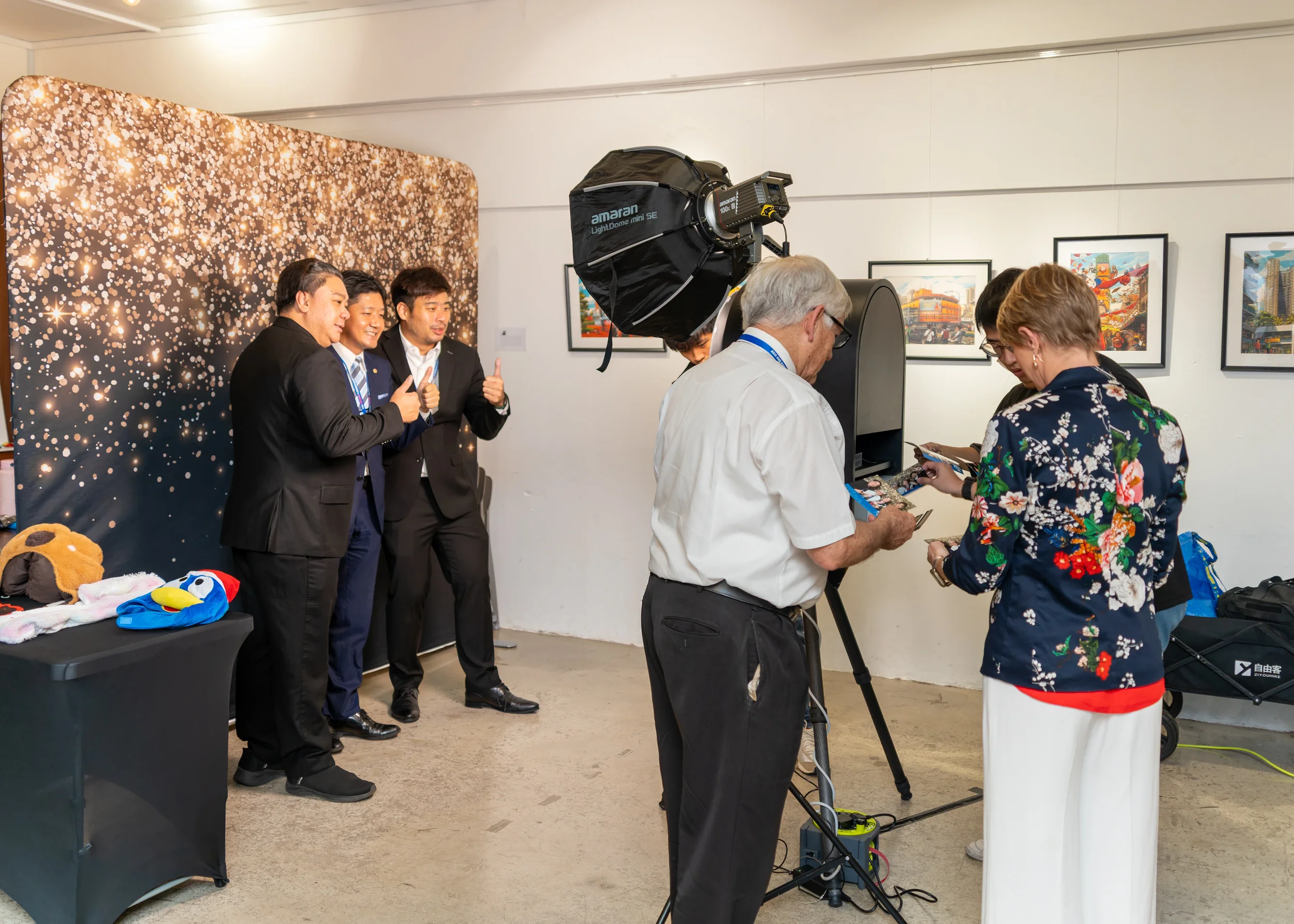 Group of people at a photo booth, some in formal attire, with a golden bokeh backdrop and framed artwork on the wall, capturing a moment during an event.