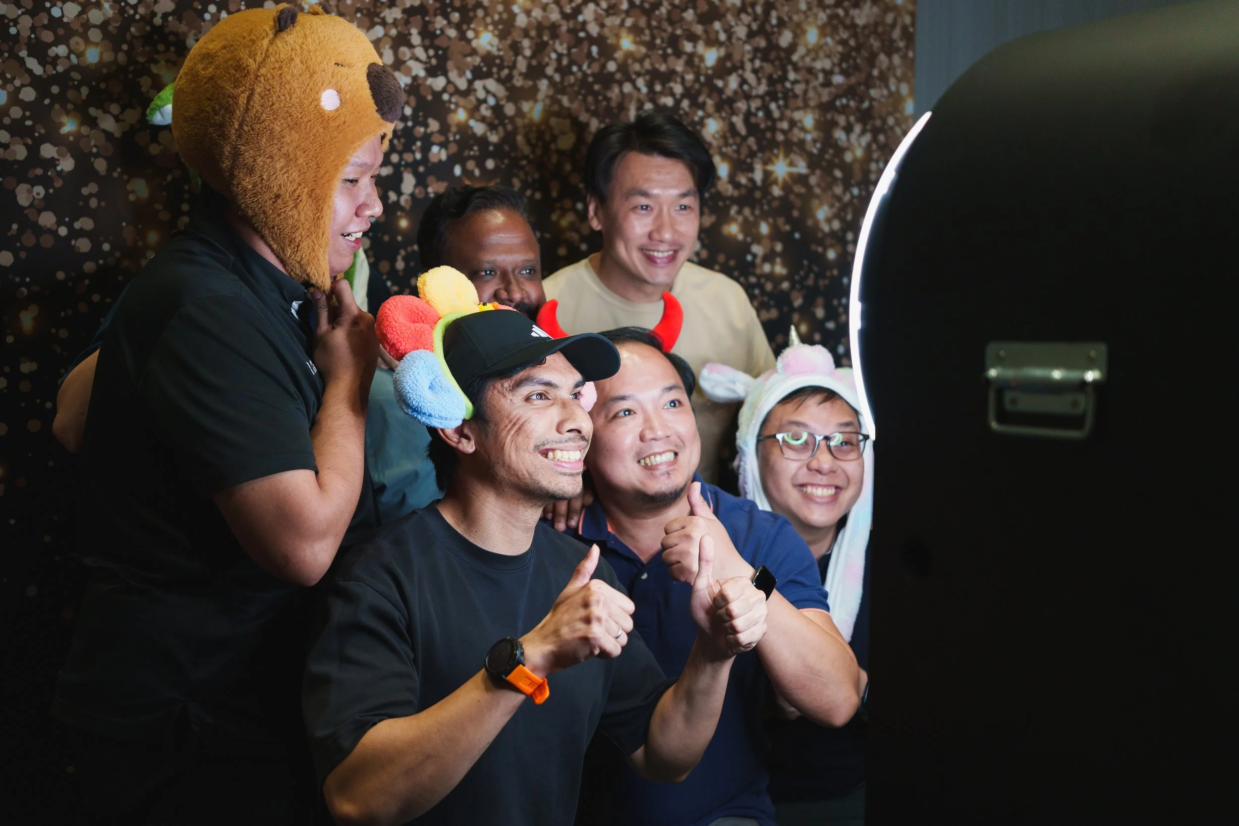 Group of friends celebrating at a photo booth, wearing colorful hats and costumes, smiling and taking a picture with a large black photo booth camera.