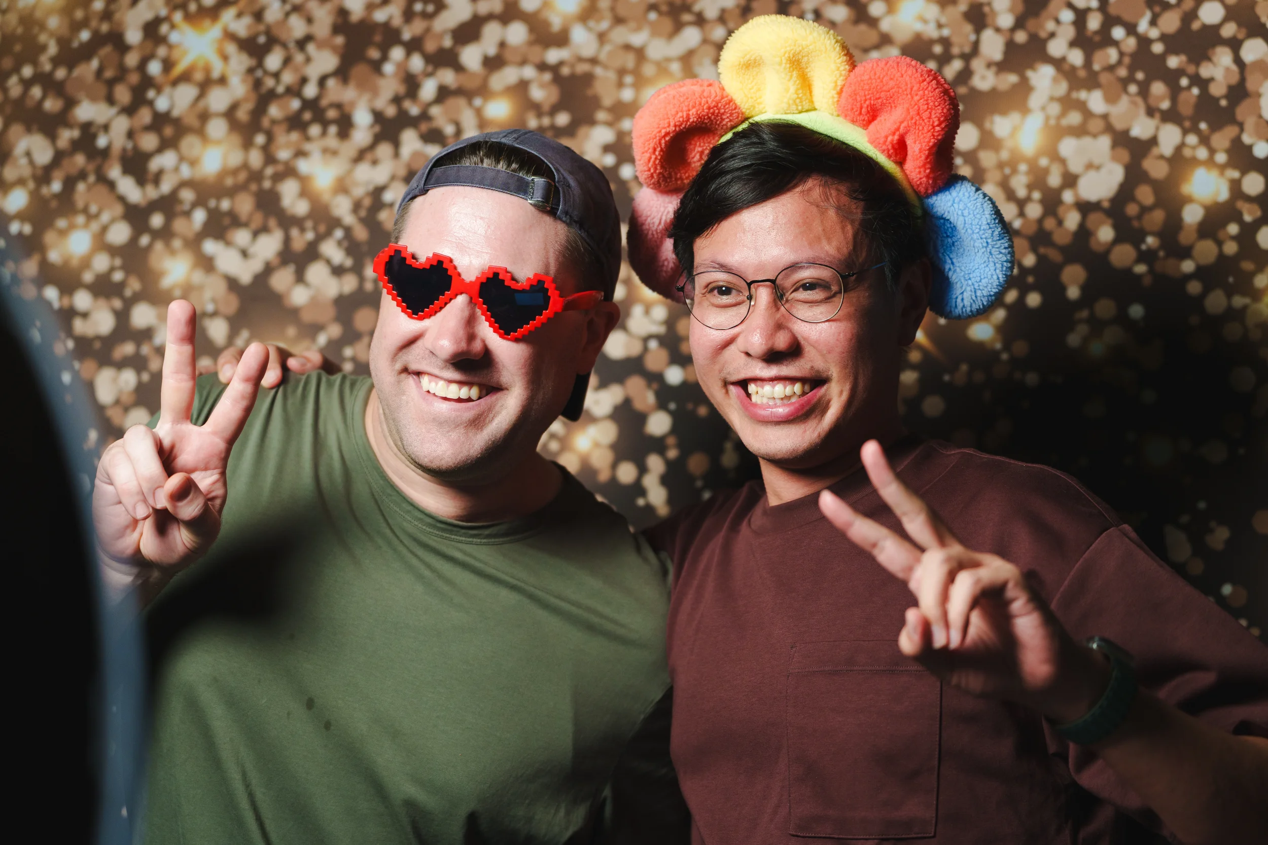 Two men smiling and making peace signs, wearing festive accessories, against a background of bokeh lights.