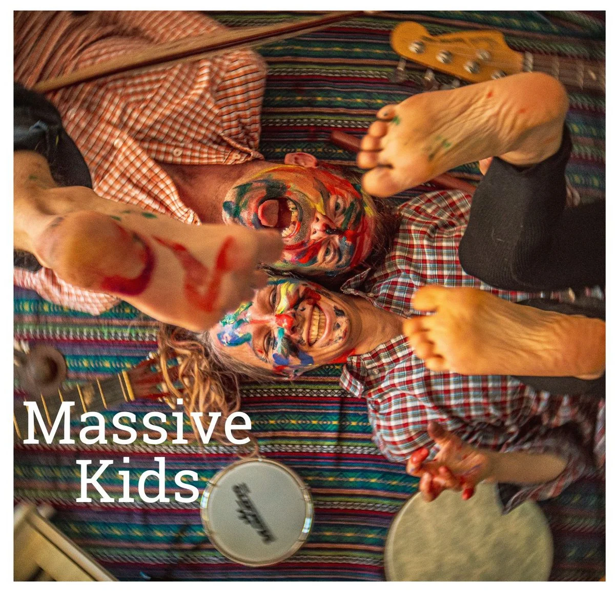 Two kids with face paint playing instruments on a colorful striped blanket. One is holding drumsticks and the other is holding a tambourine. The text 'Massive Kids' is overlaid on the image.