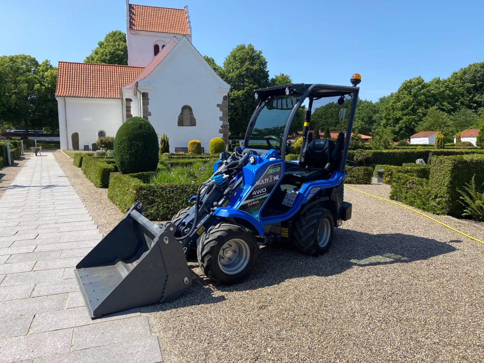 En MultiOne är parkerad på en grusplan framför en vit stenbyggnad, troligen en kyrka. Vägen till vänster är stenlagd och det är gröna träd och buskar runt omkring.