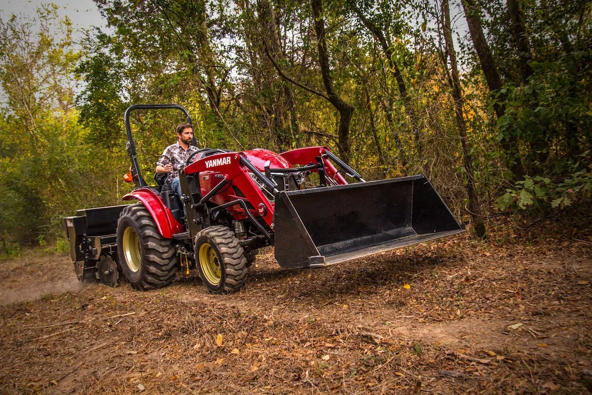 Person som kör en röd Yanmar-tractor med skopa framför i ett skogsområde med lövträd.