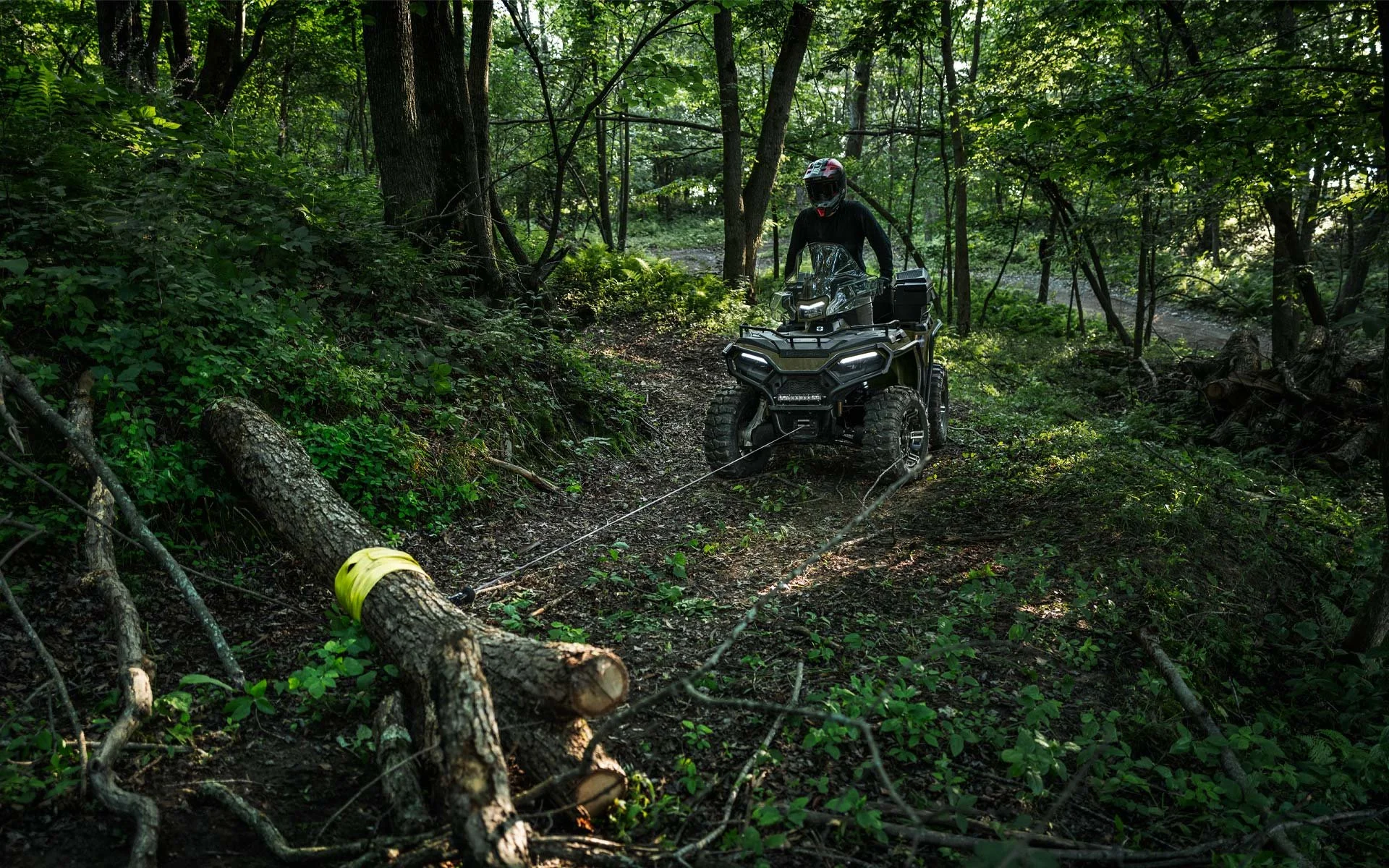 En person på en Polaris Sportsman 570 kör genom en tät skog med gröna träd och buskar. Fyrhjulingen är svart och har starka däck, och personen är iklädd hjälm och mörka kläder.