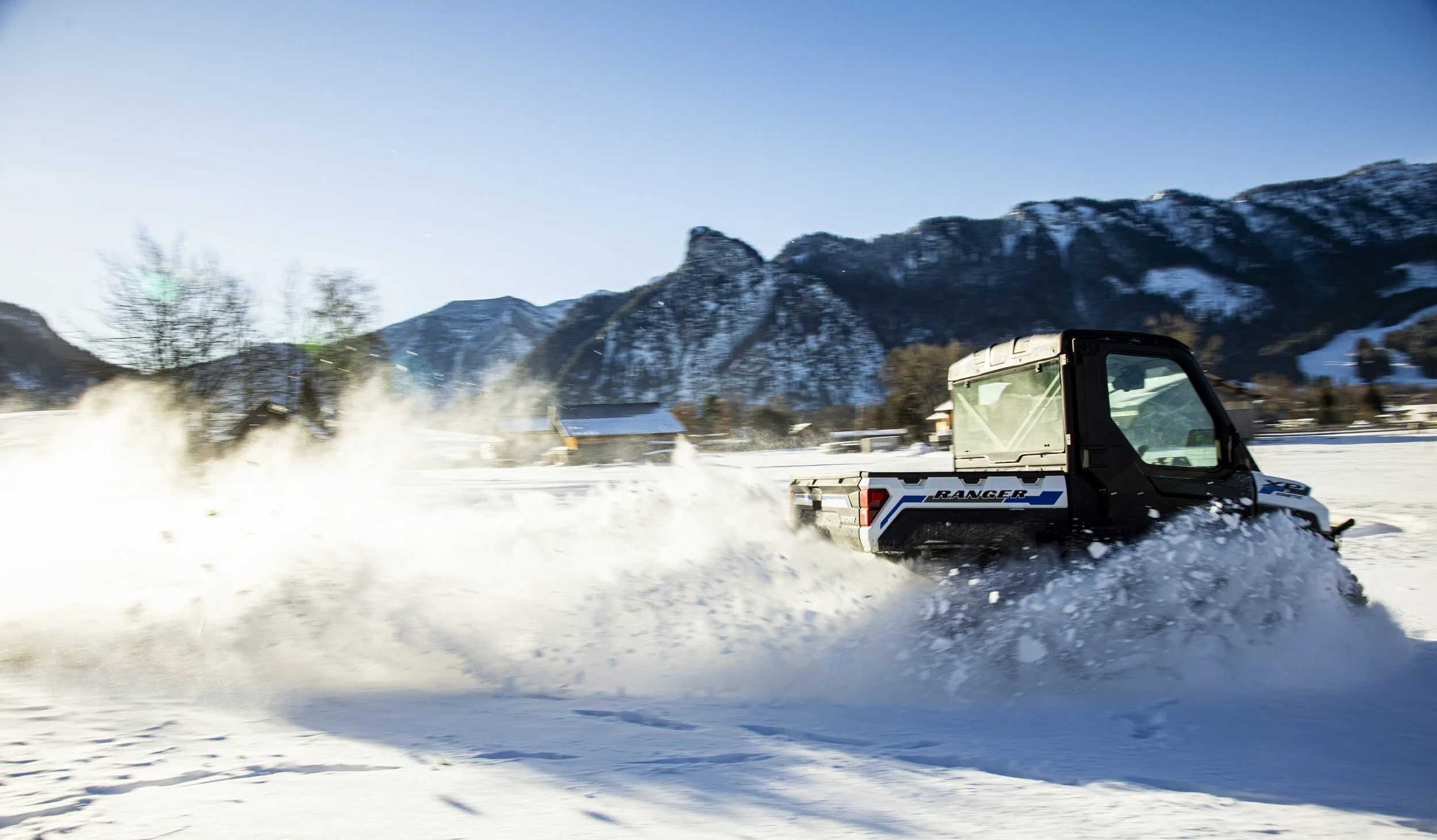 En Polaris Ranger XP Kinetic som kör genom snö, med snöbergen i bakgrunden under en klar blå himmel.