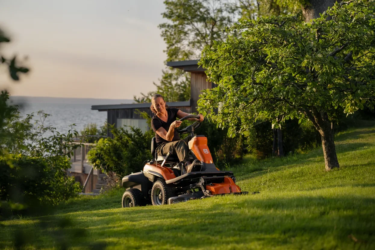 Kvinna klipptrimmar gräset med en Husqvarna Rider i en trädgård med träd och ett hus i bakgrunden under solnedgången.