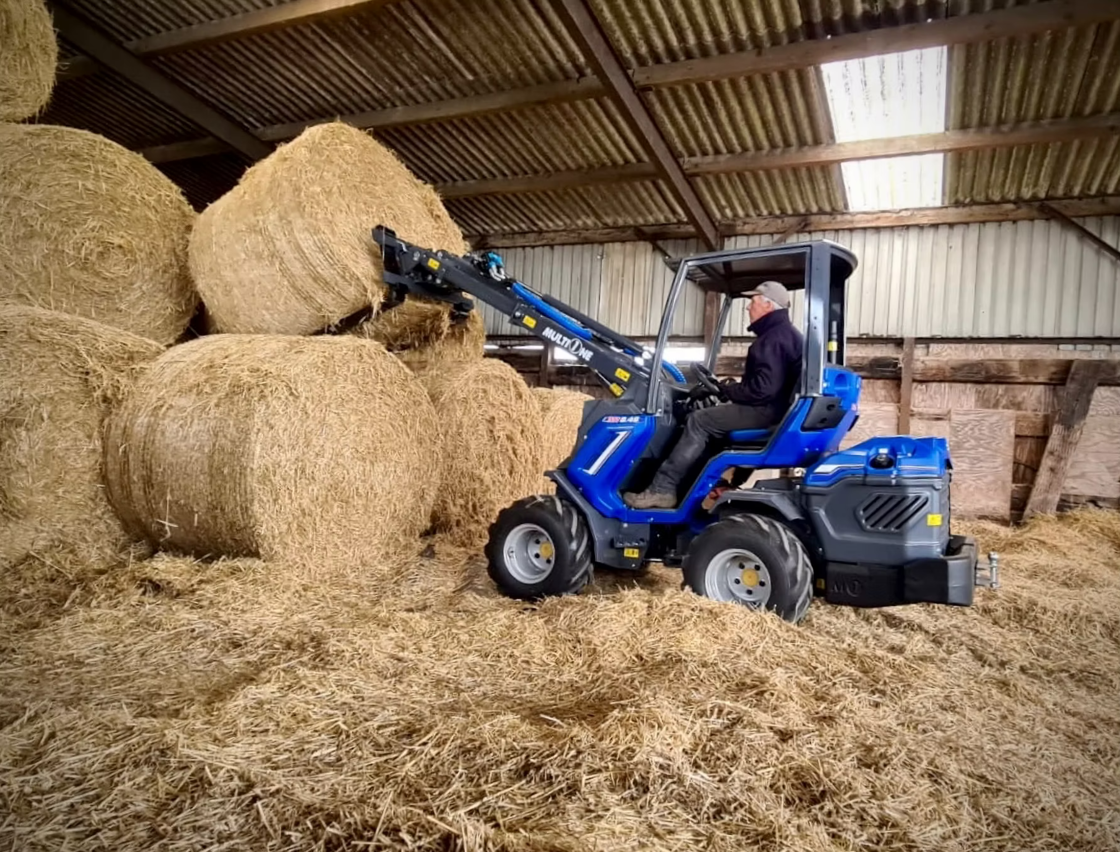 En person kör en MultiOne med ett utskjutande armverktyg inuti ett förråd med höbalar och halm.
