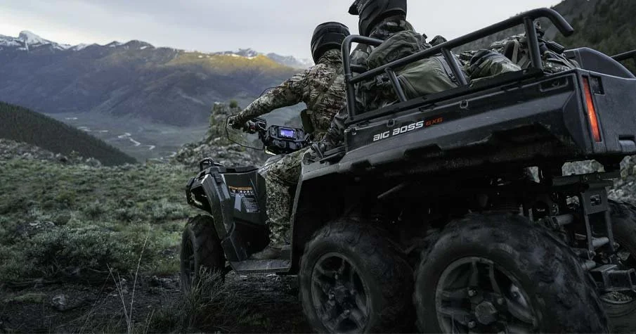 Två personer i militäruniformer och hjälmar som kör en Polaris Sportsman 570 6x6 genom ett bergsområde med gröna buskar och berg i bakgrunden.