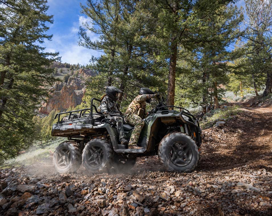 Två personer i hjälmar kör en Polaris Sportsman 570 6x6 på en stig i en skog med tallar och berg i bakgrunden.