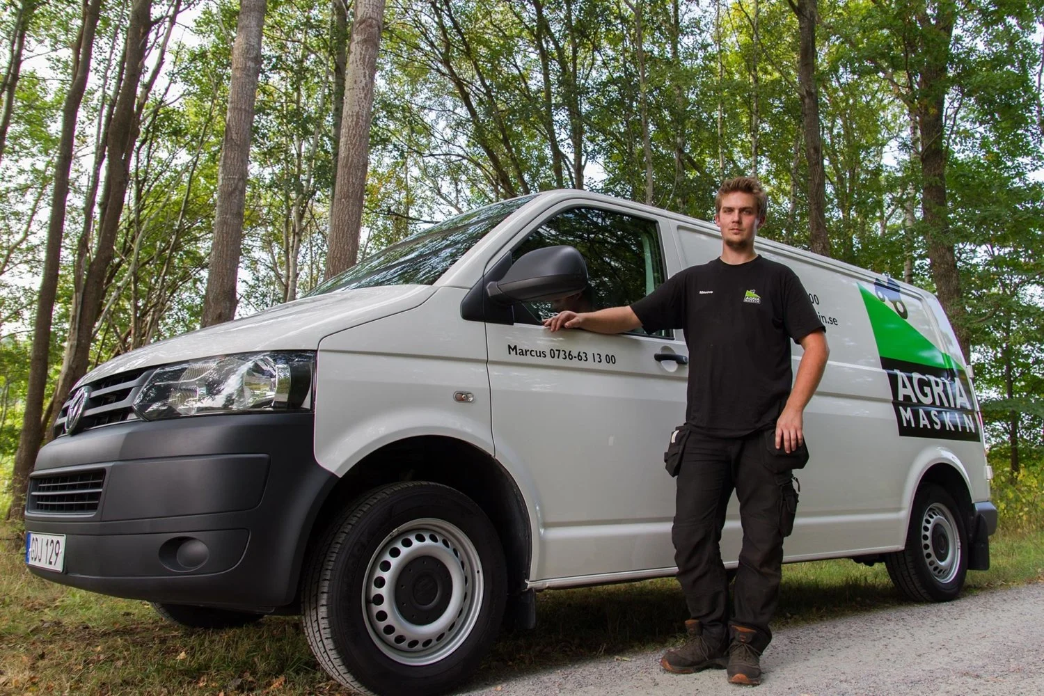 En ung man står bredvid en vit företagsbil i en skogsmiljö. Mannen bär en svart t-shirt och arbetsbyxor. Bilen har text och kontaktuppgifter på sidan, inklusive namn och telefonnummer.